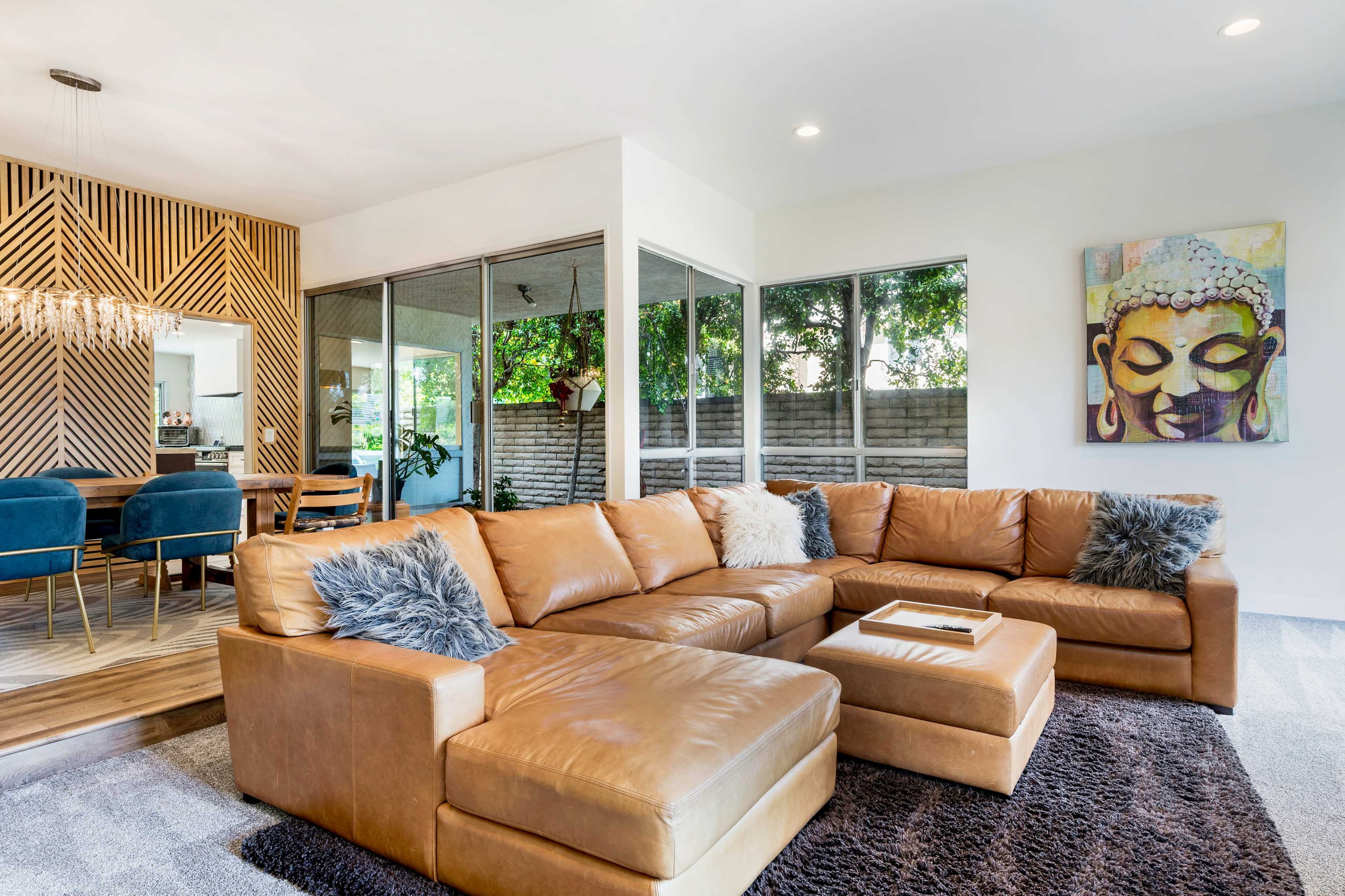 The image shows a spacious living room with a large brown leather sectional sofa, a dark area rug, a dining area in the background, and a decorative Buddha painting on the wall.