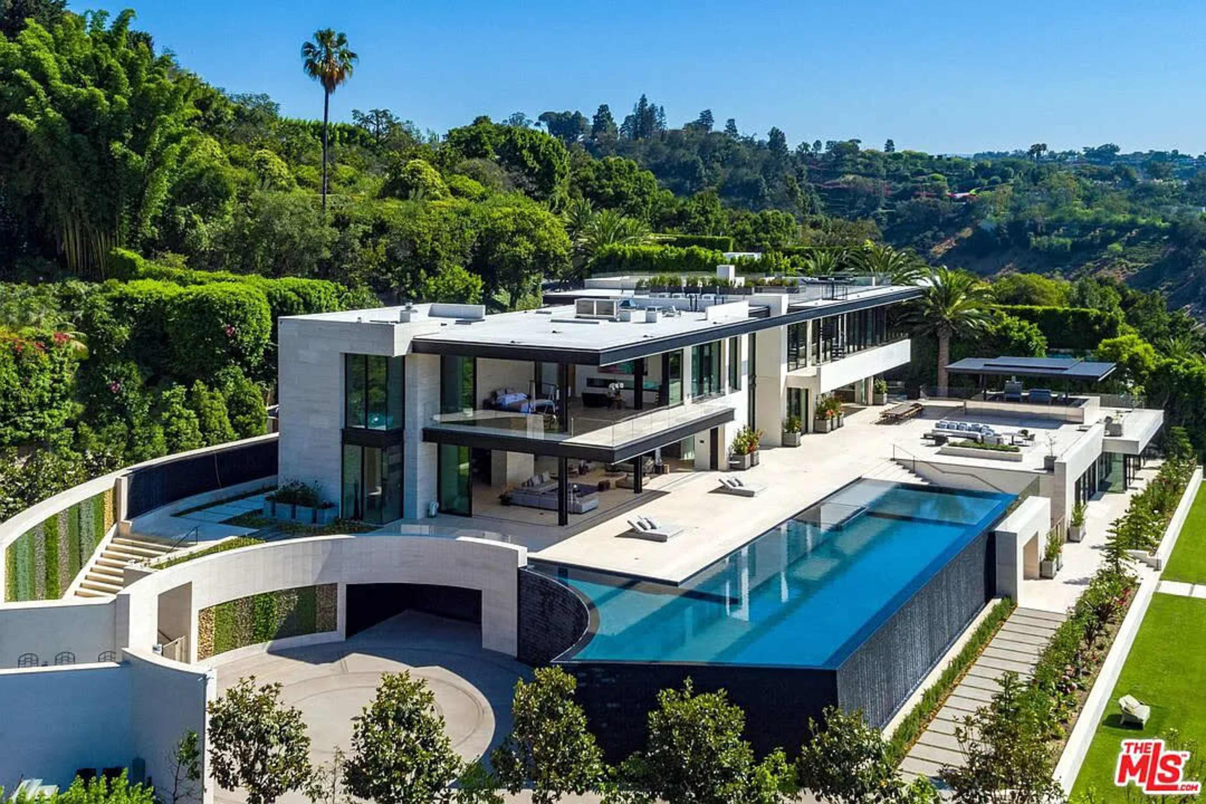 A large modern mansion features multiple levels, expansive terraces, and a swimming pool surrounded by landscaped gardens.