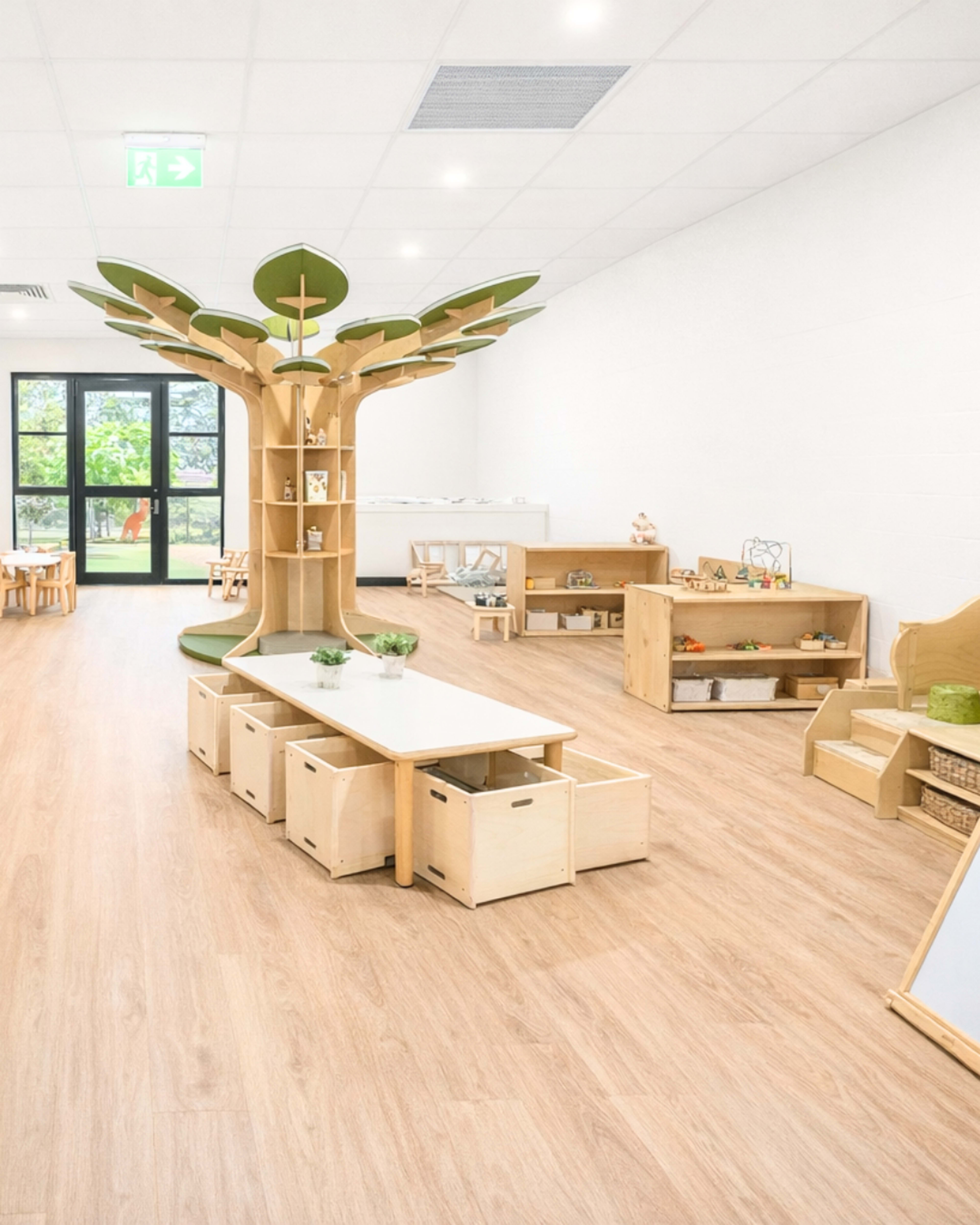 The image shows a bright classroom designed for children, featuring a wooden tree structure, tables, and various storage units arranged in a spacious layout.