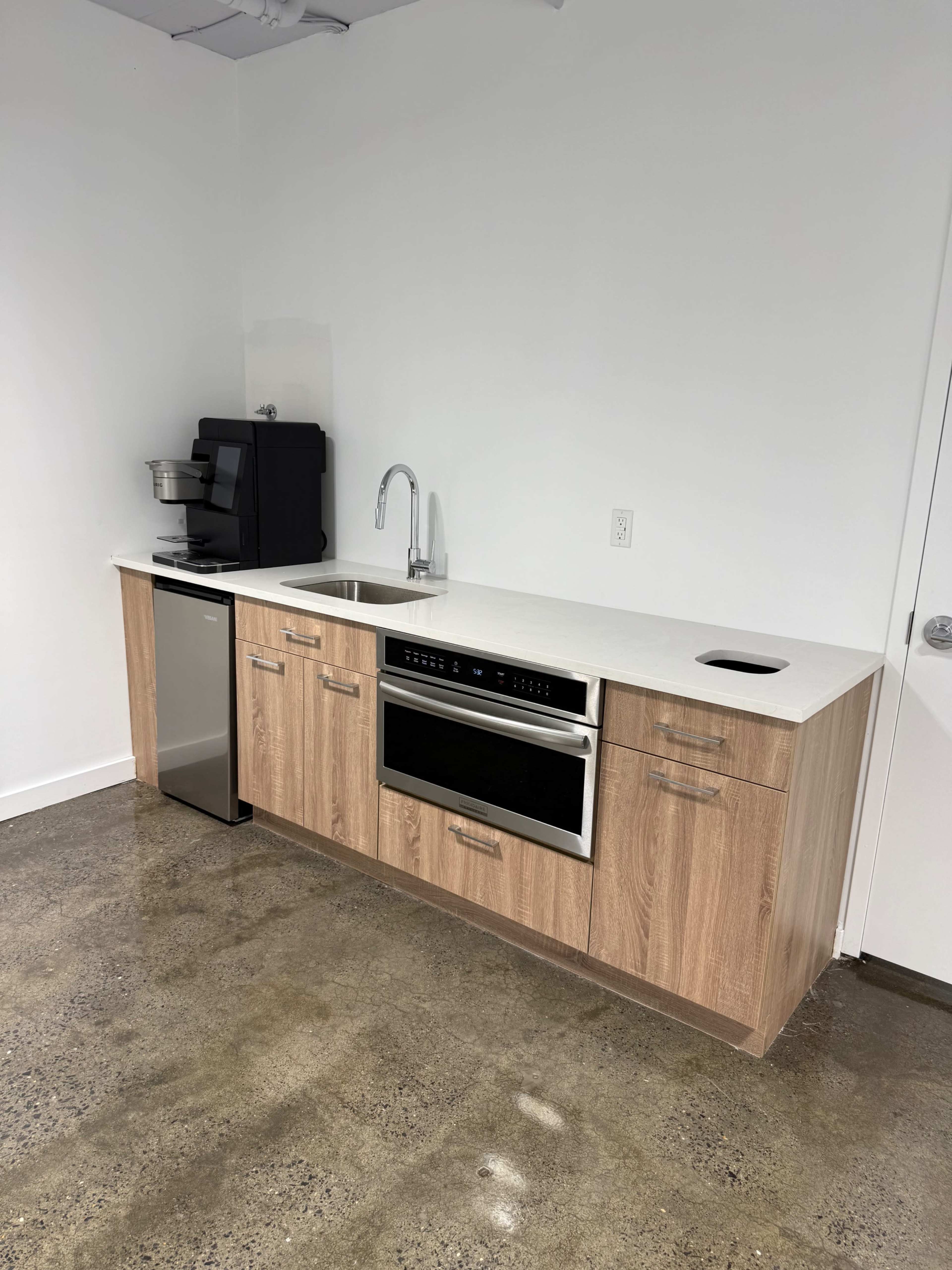 The image shows a modern kitchen setup featuring a countertop with a sink, an oven, a coffee maker, and a refrigerator, all installed in a well-lit space with a concrete floor.