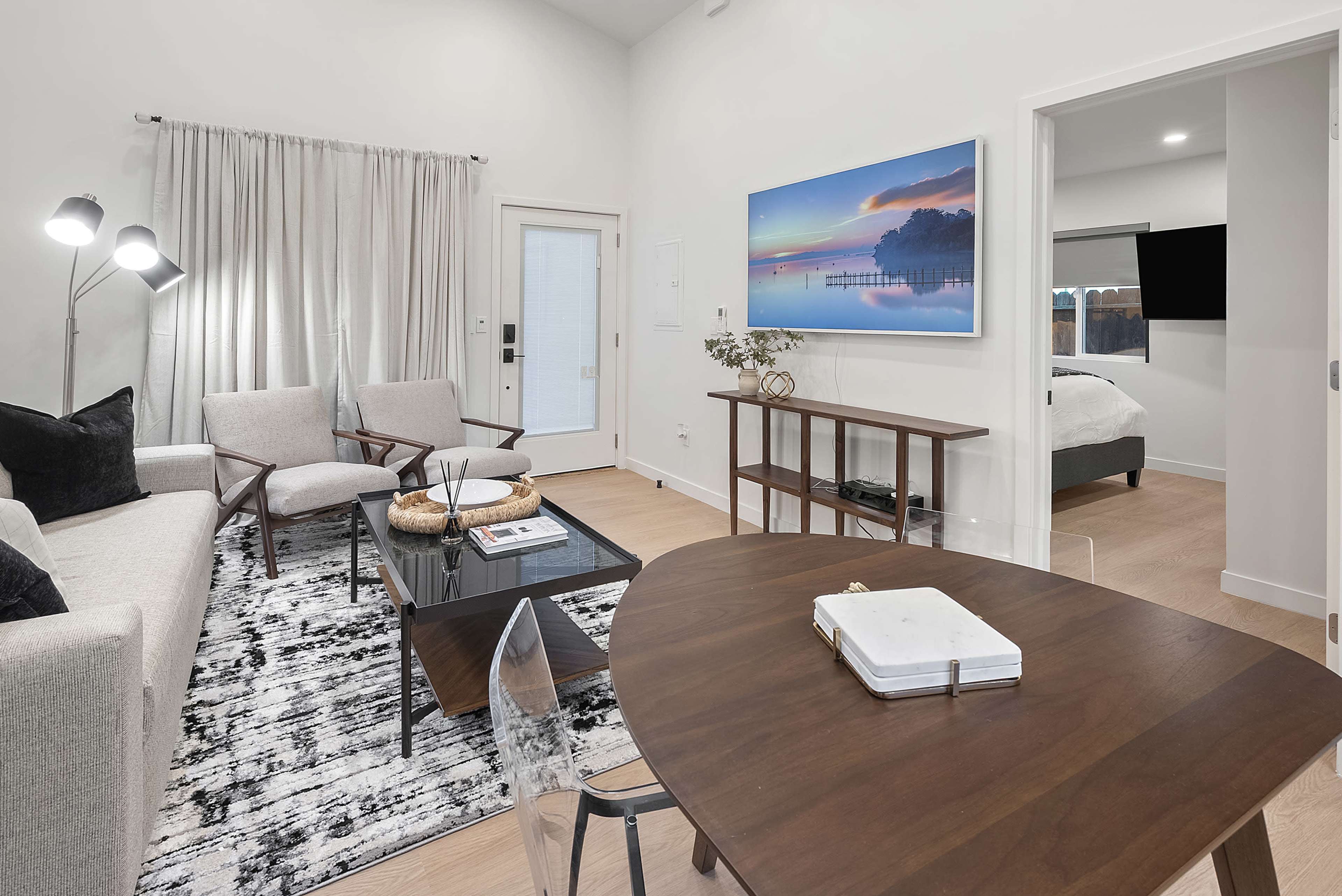 The image shows a modern living area with a gray sofa, two chairs, a coffee table, and a television mounted on the wall, all set against a backdrop of light-colored walls and wooden flooring.