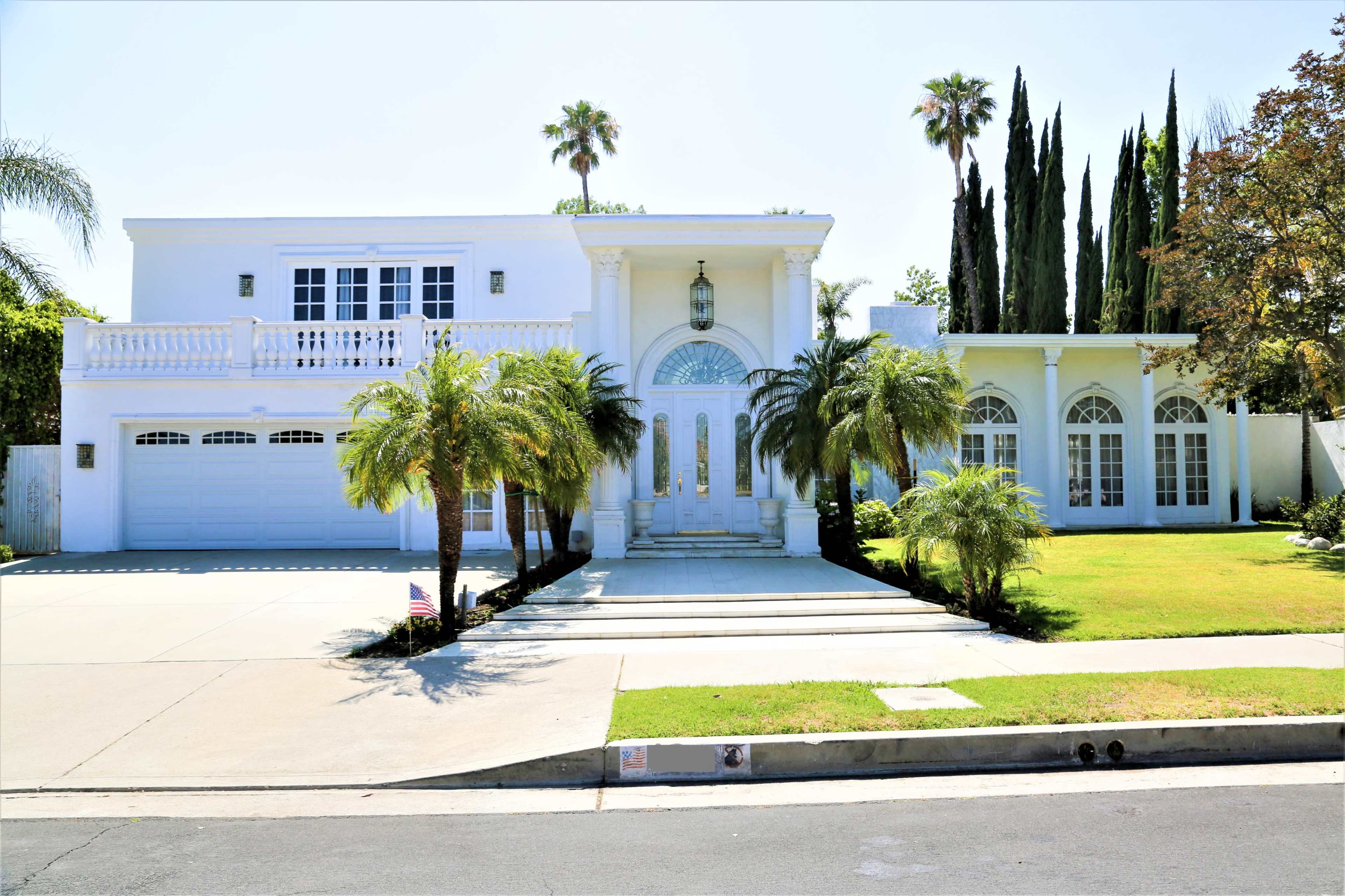 A large white house with a grand entrance, palm trees, and a well-maintained lawn is set at the end of a driveway.