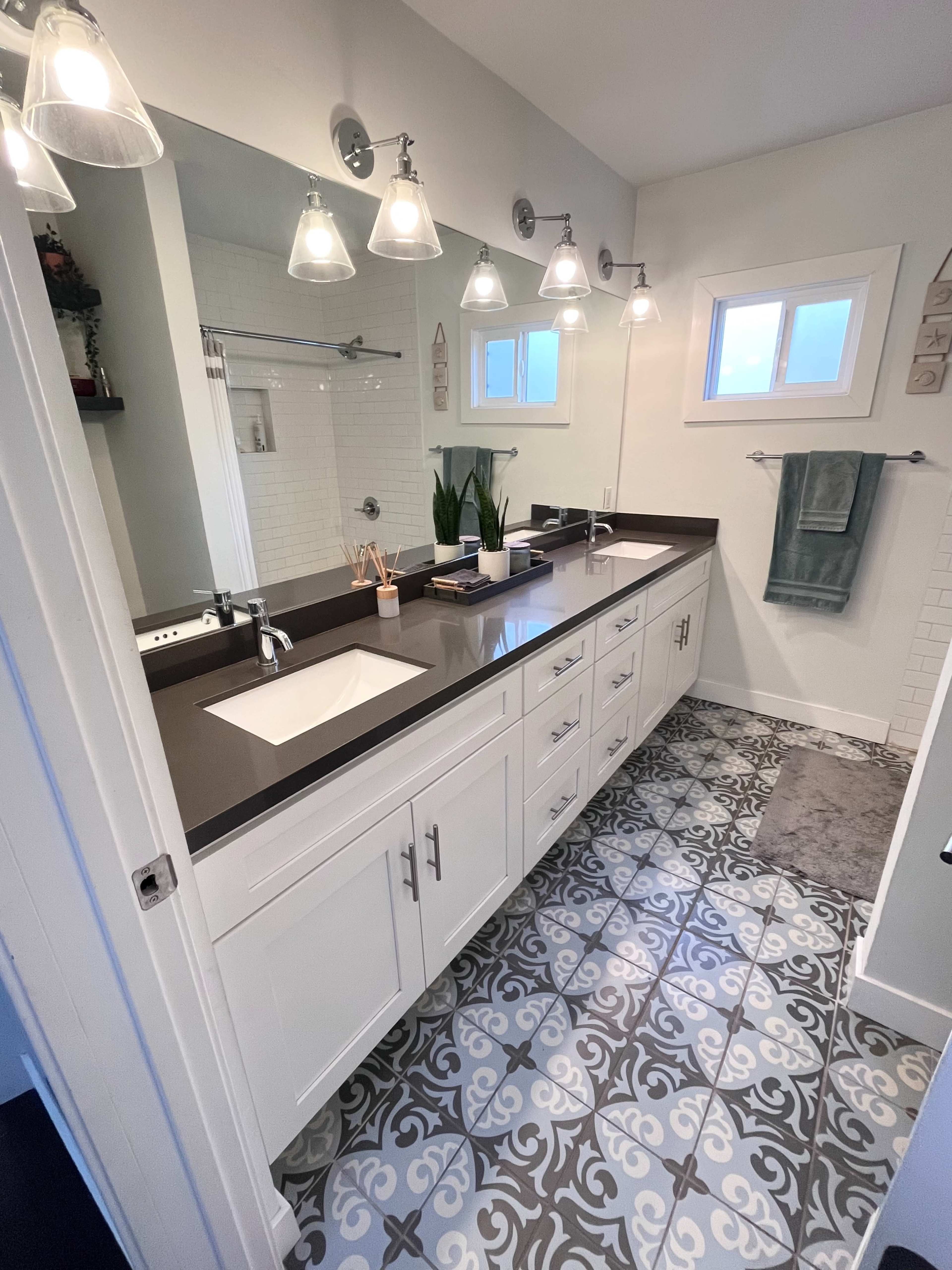 A modern bathroom with a double vanity, light fixtures above the mirrors, and patterned tiled flooring.