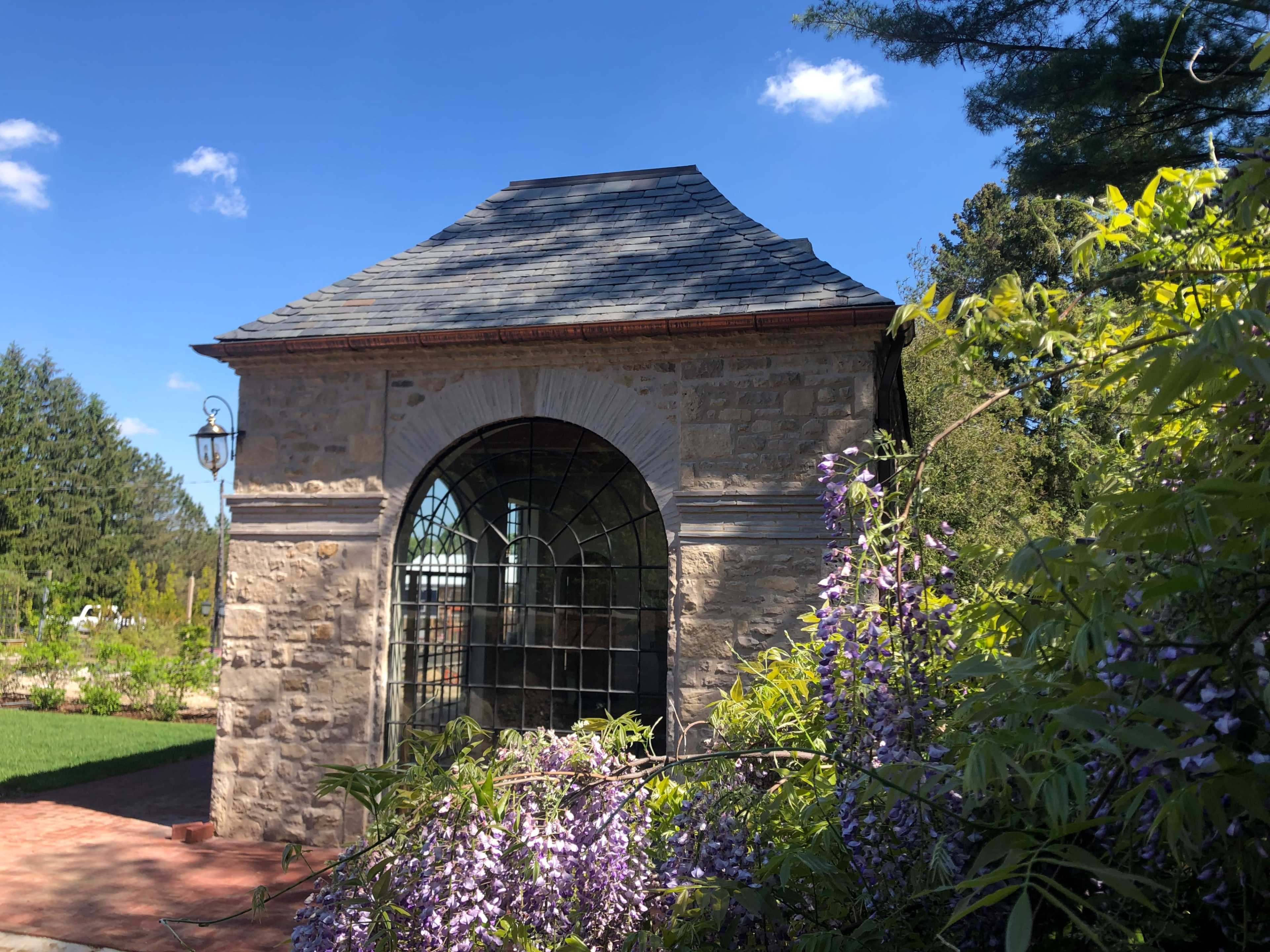 A stone gazebo with a sloped roof and a large arched window, surrounded by purple flowering plants and trees under a blue sky.