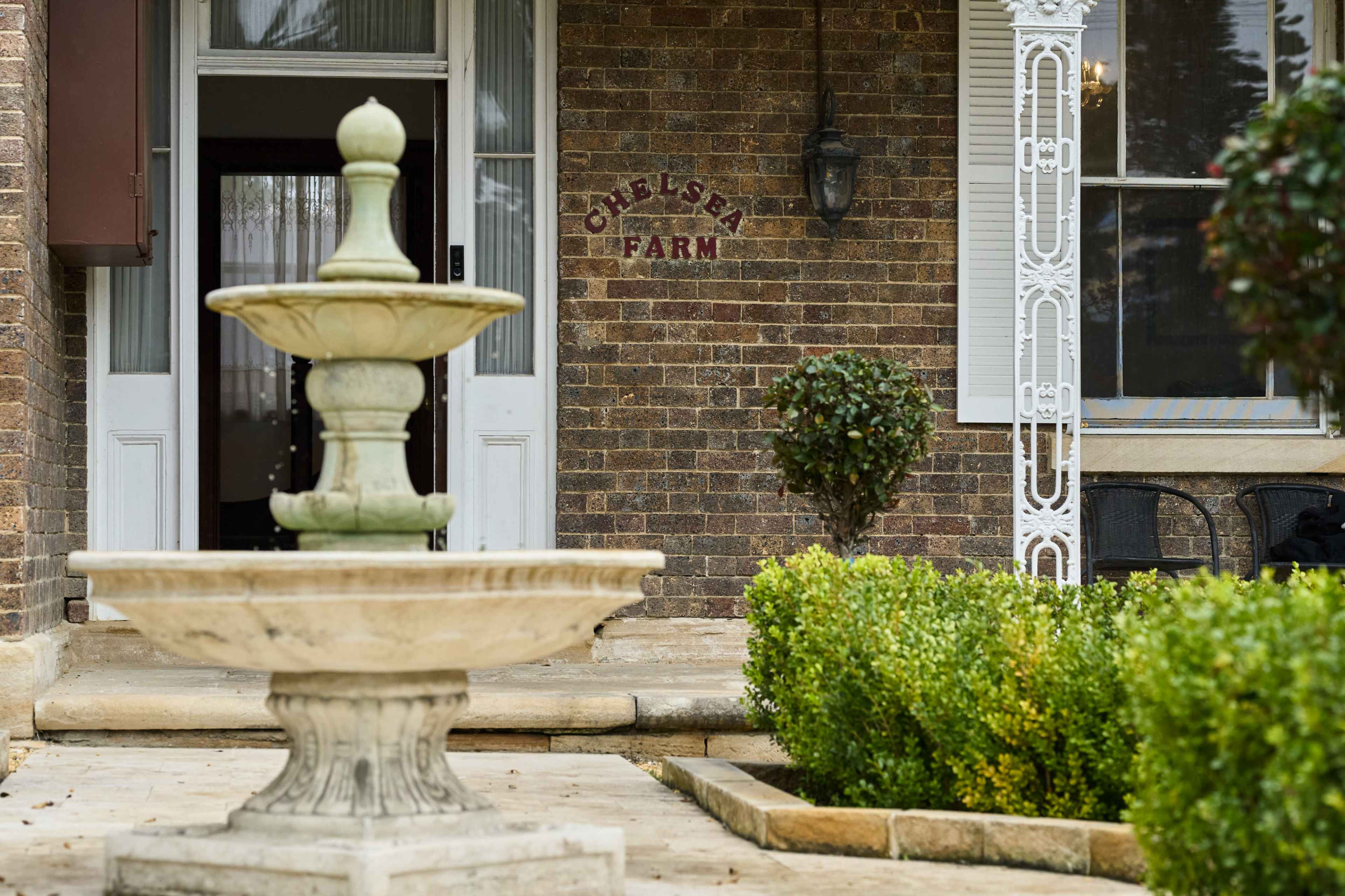 The image shows a brick facade of a building with the words "Chelsea Farm" displayed, featuring a stone fountain in the foreground and neatly trimmed hedges.