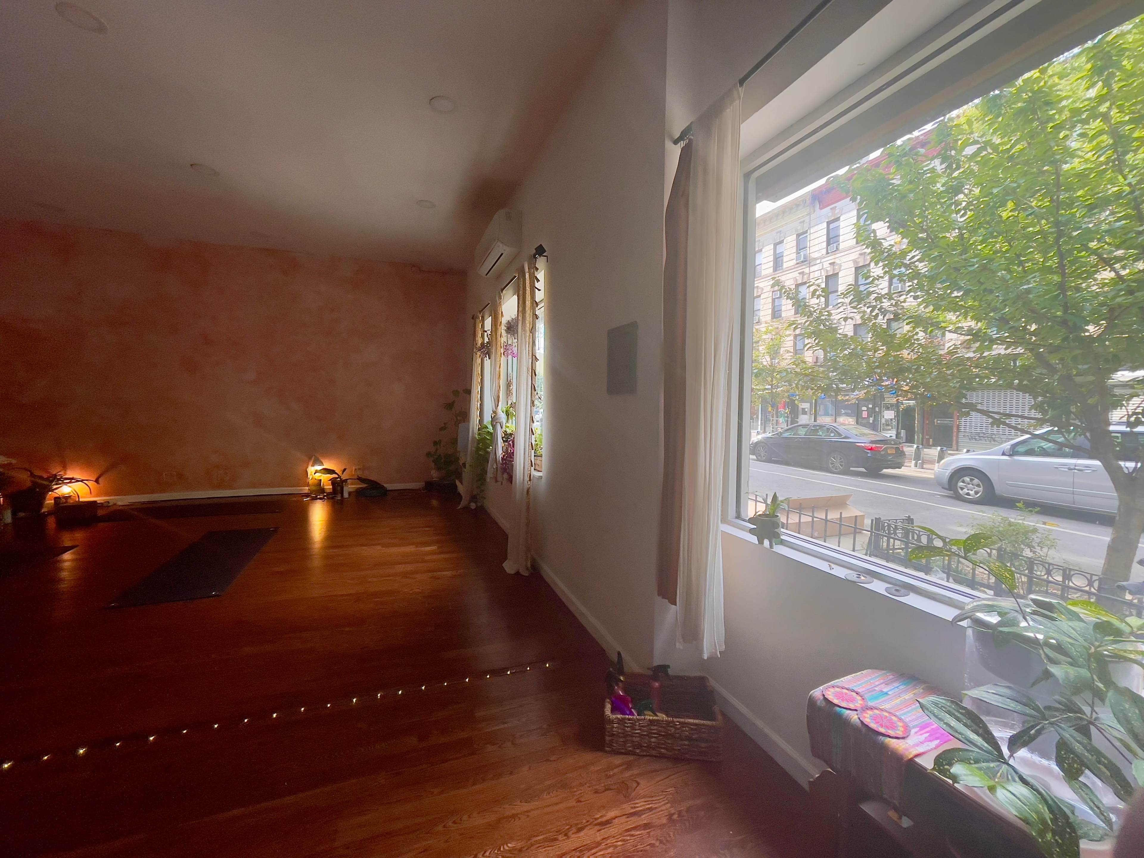 The image shows a spacious yoga studio with wooden floors, large windows allowing natural light, and a view of a street outside.