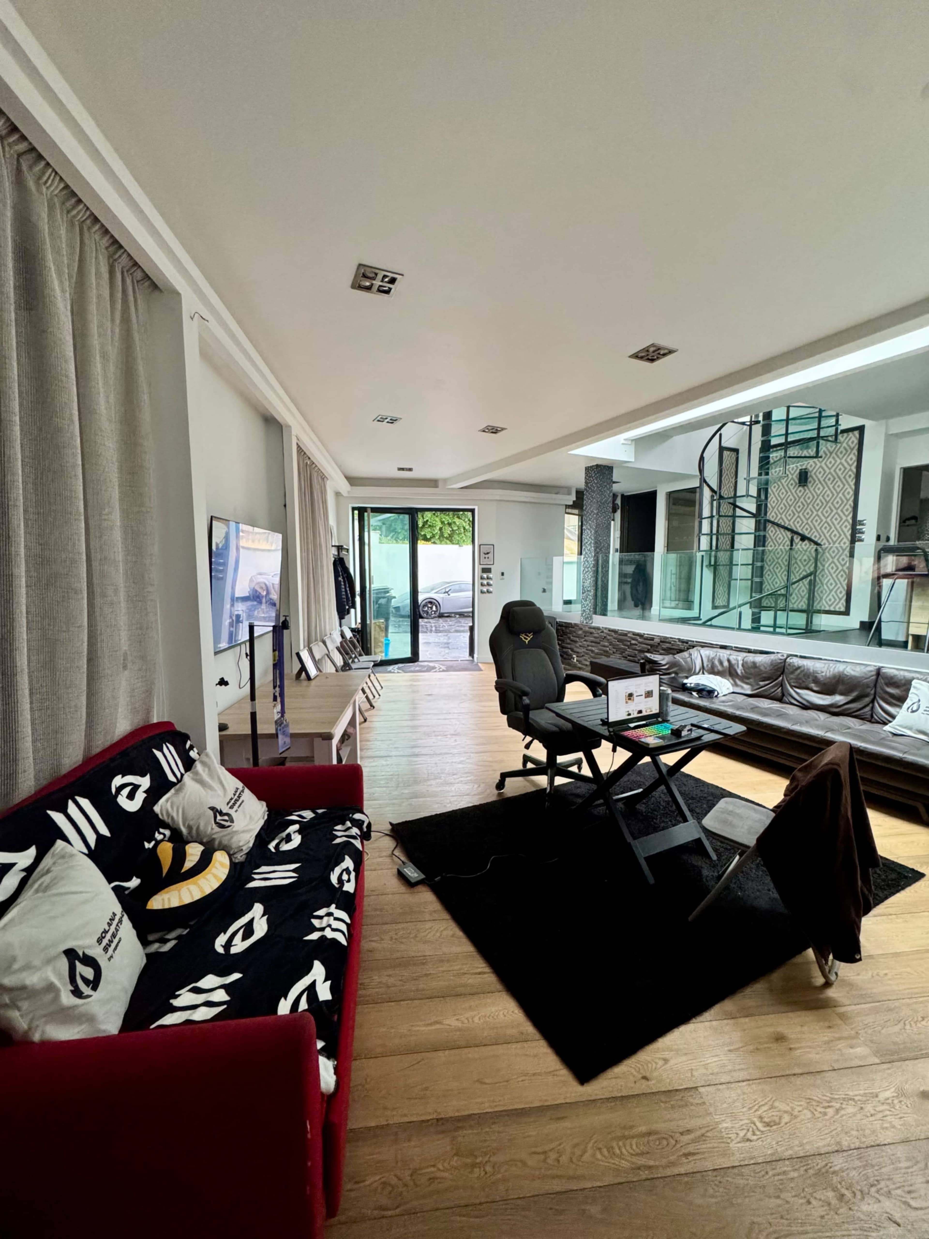The image depicts a modern living room with a red couch, a computer chair at a desk, a large glass door leading outside, and a spiral staircase in the background.