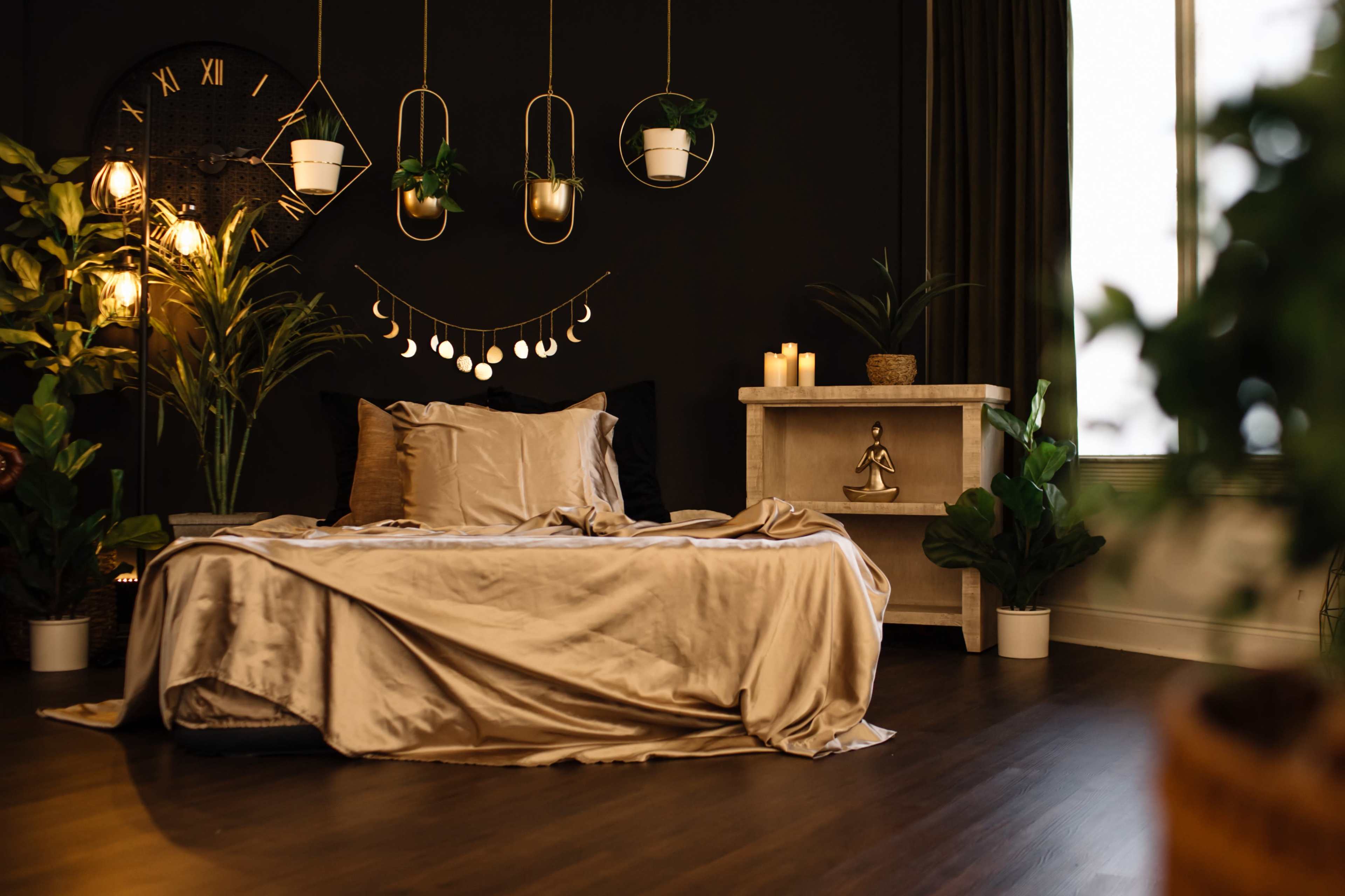 The image shows a cozy bedroom with a dark wall, a large clock, plants in hanging pots, and a neatly made bed with a satin beige duvet.