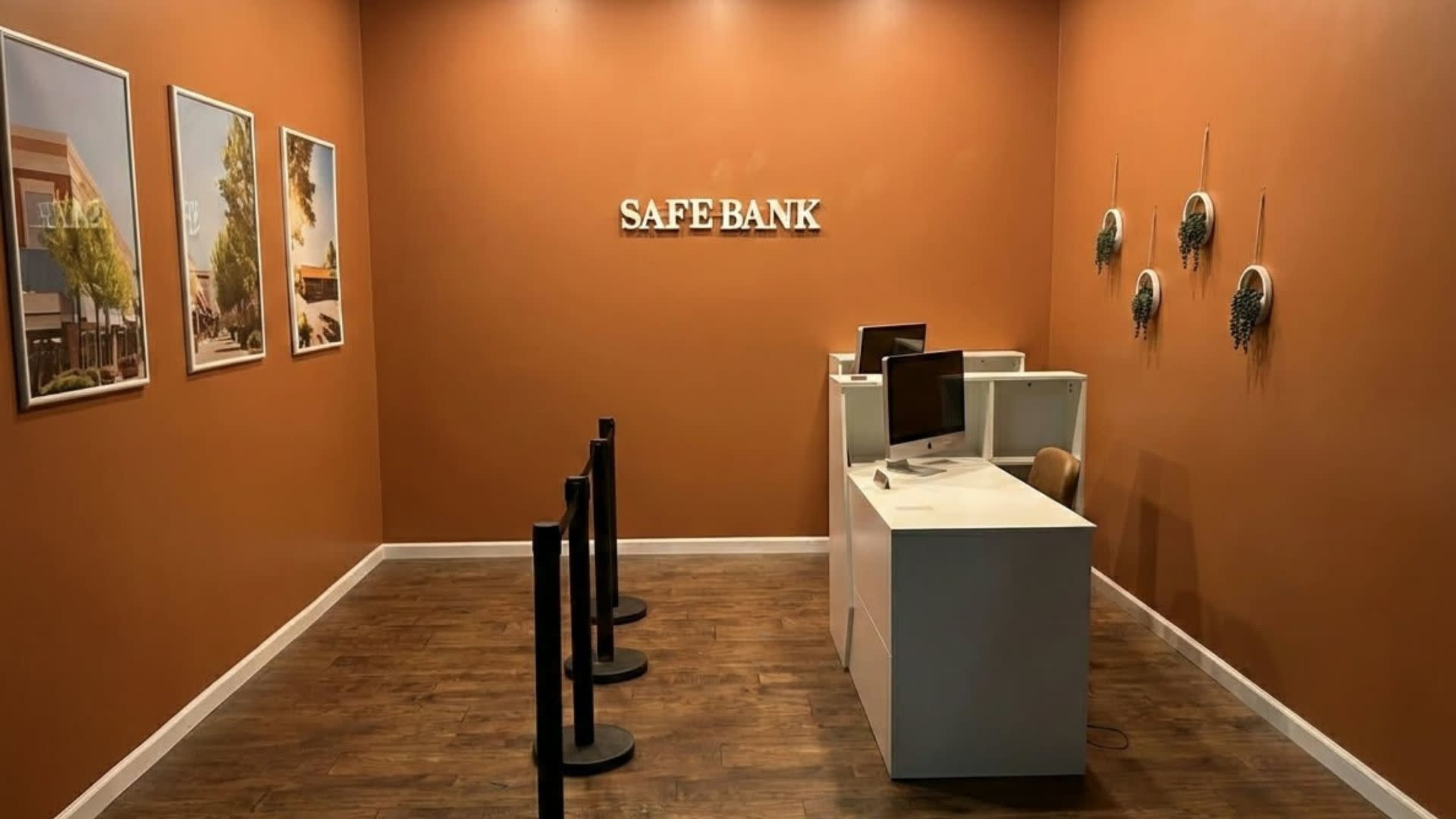 The image shows a bank interior with an orange wall, two workstations with computers, and a queue management system alongside framed pictures on the wall.