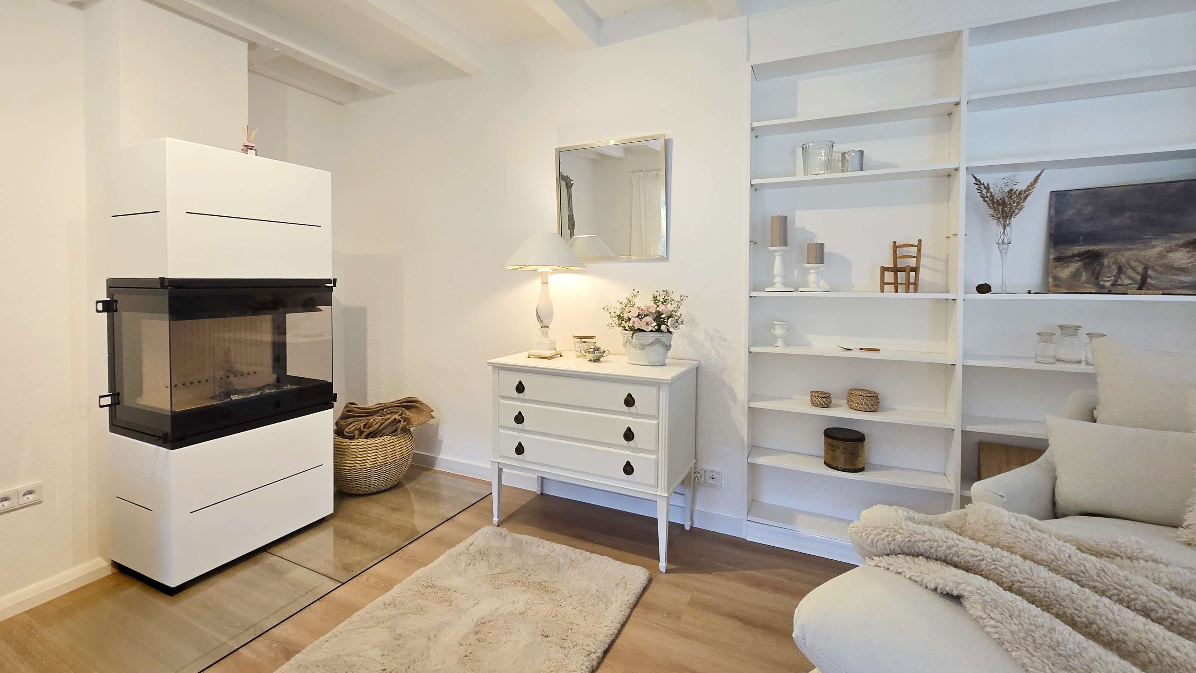 The image shows a modern living room with a white decorative fireplace, a small table with a lamp and flowers, a shelf with various decorative items, and a light-colored rug on the floor.