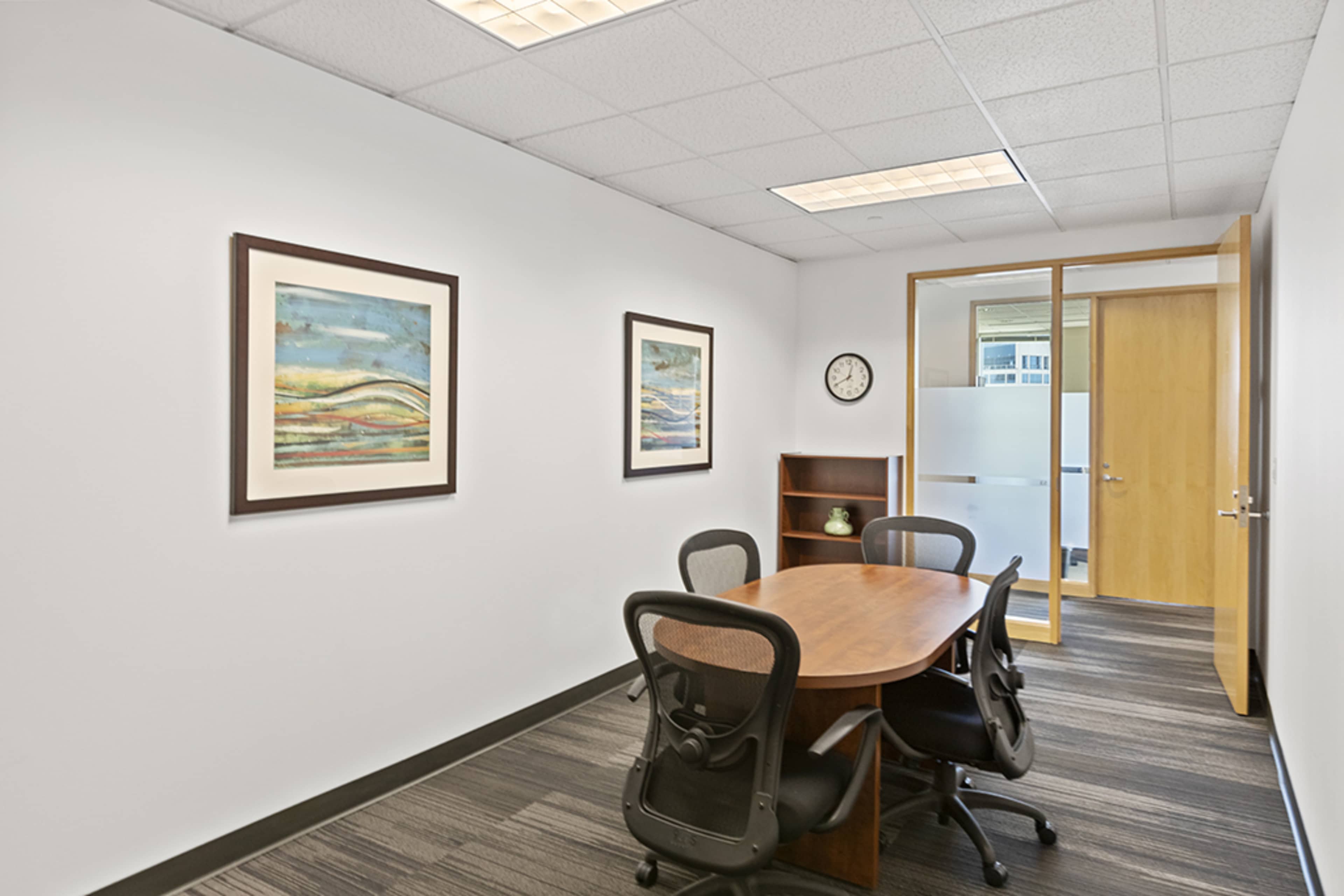 A conference room features a round wooden table surrounded by four black chairs, with two framed paintings on the white walls and a clock on the shelf.