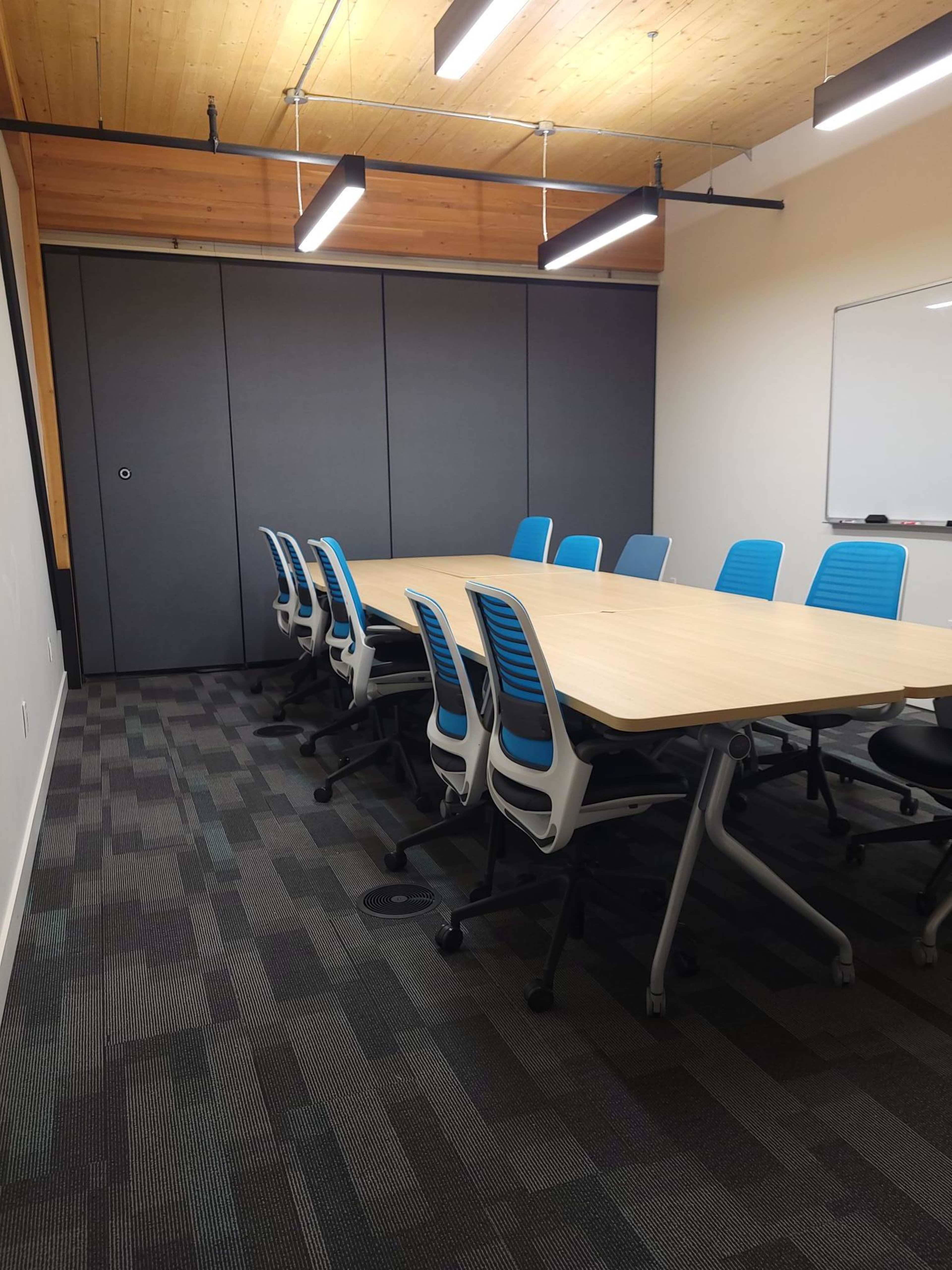 A modern conference room features a long wooden table surrounded by blue office chairs, with a whiteboard and storage cabinet in the background.