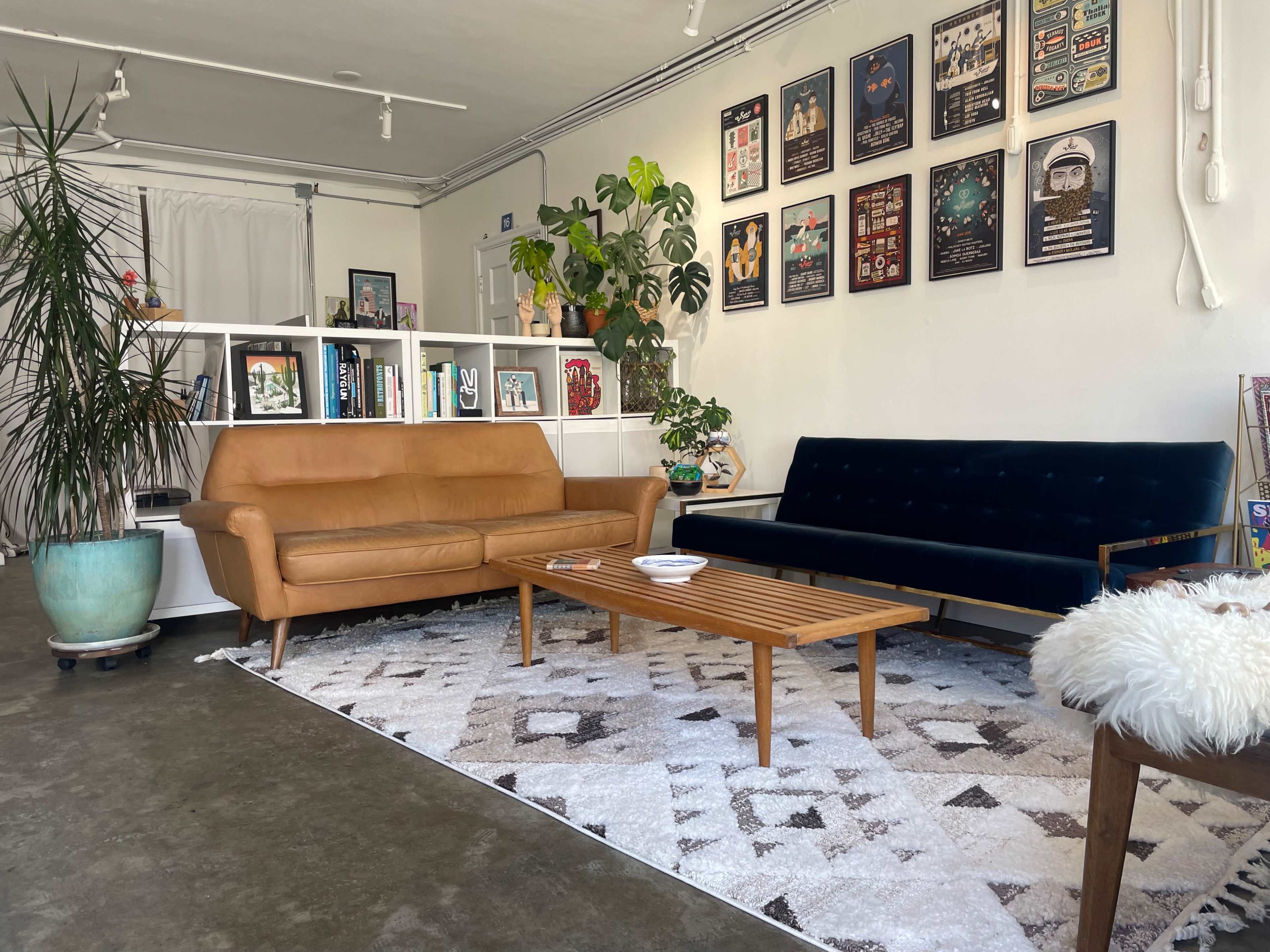 The image shows a stylish living room with a brown leather sofa, a blue velvet couch, a wooden coffee table, and a rug, all arranged around a wall featuring framed artwork.