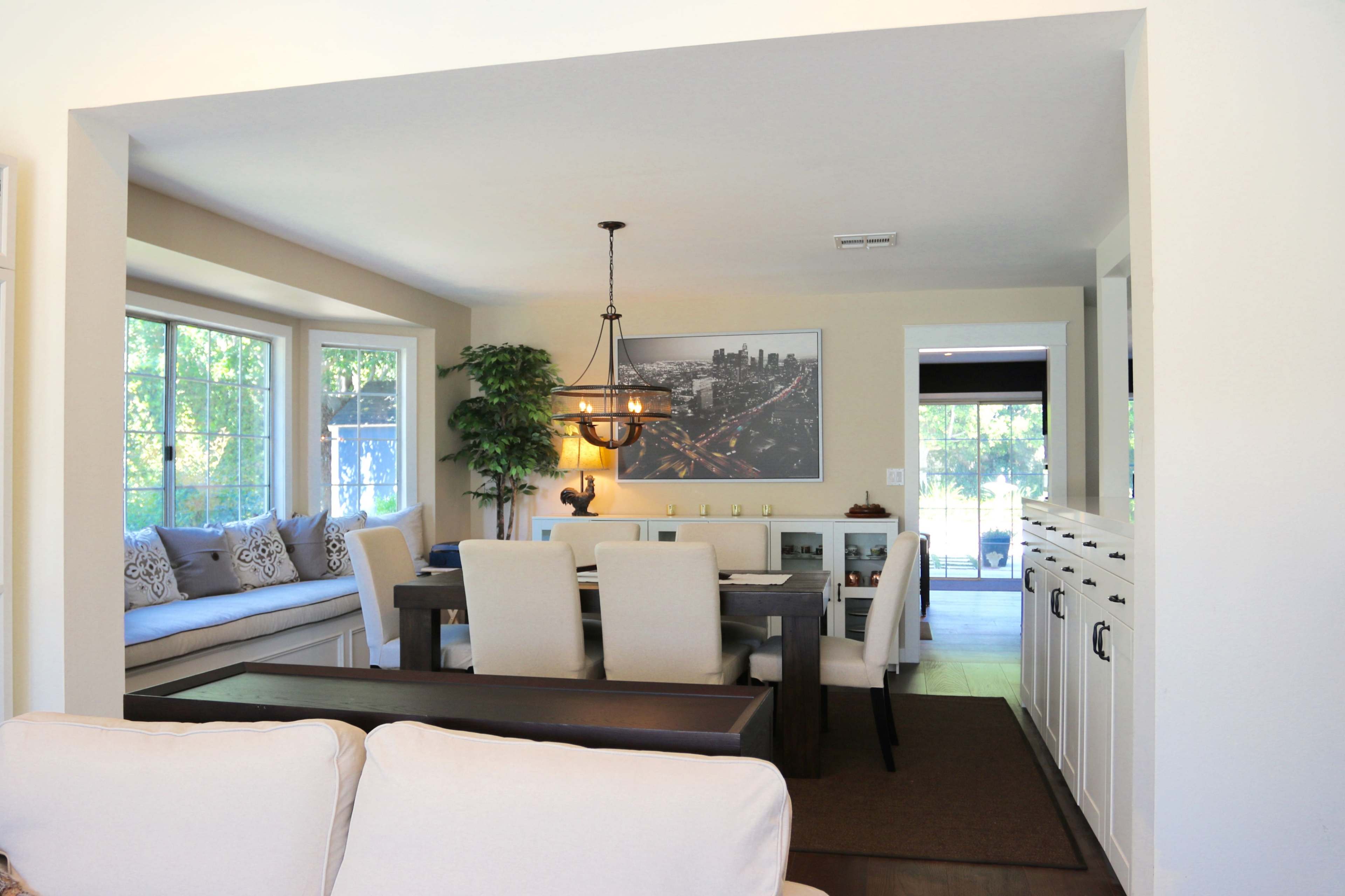A spacious dining area features a wooden table surrounded by beige chairs, with a large window and a cozy seating nook visible.