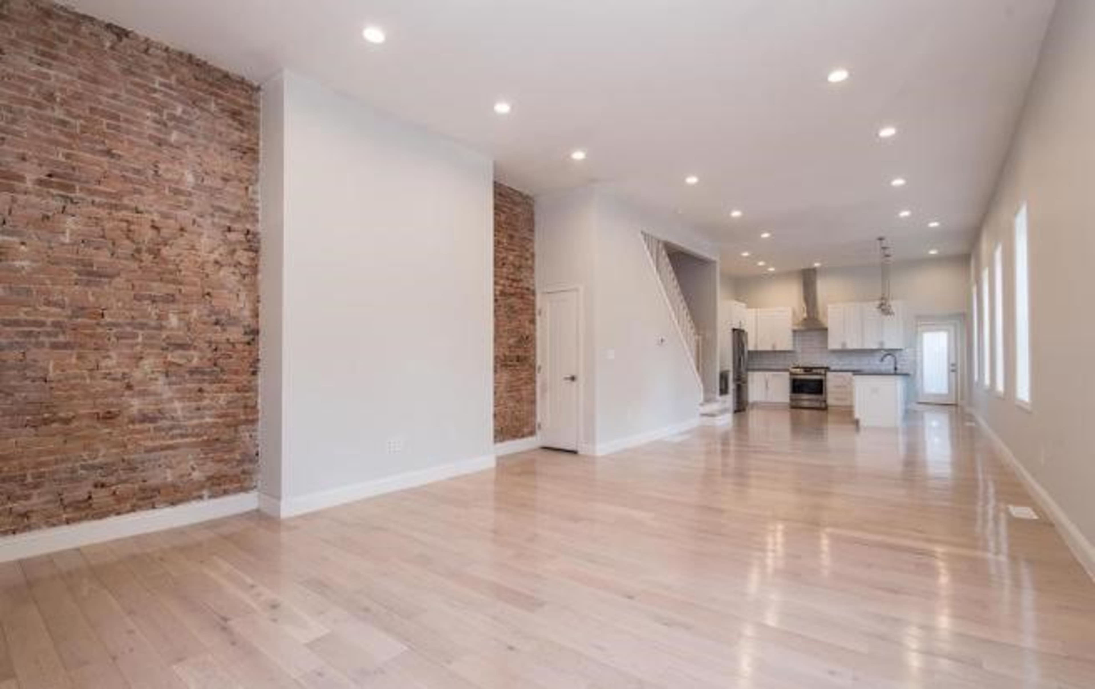 A spacious interior with a mix of exposed brick and smooth white walls, hardwood floors, and a kitchen area visible in the background.