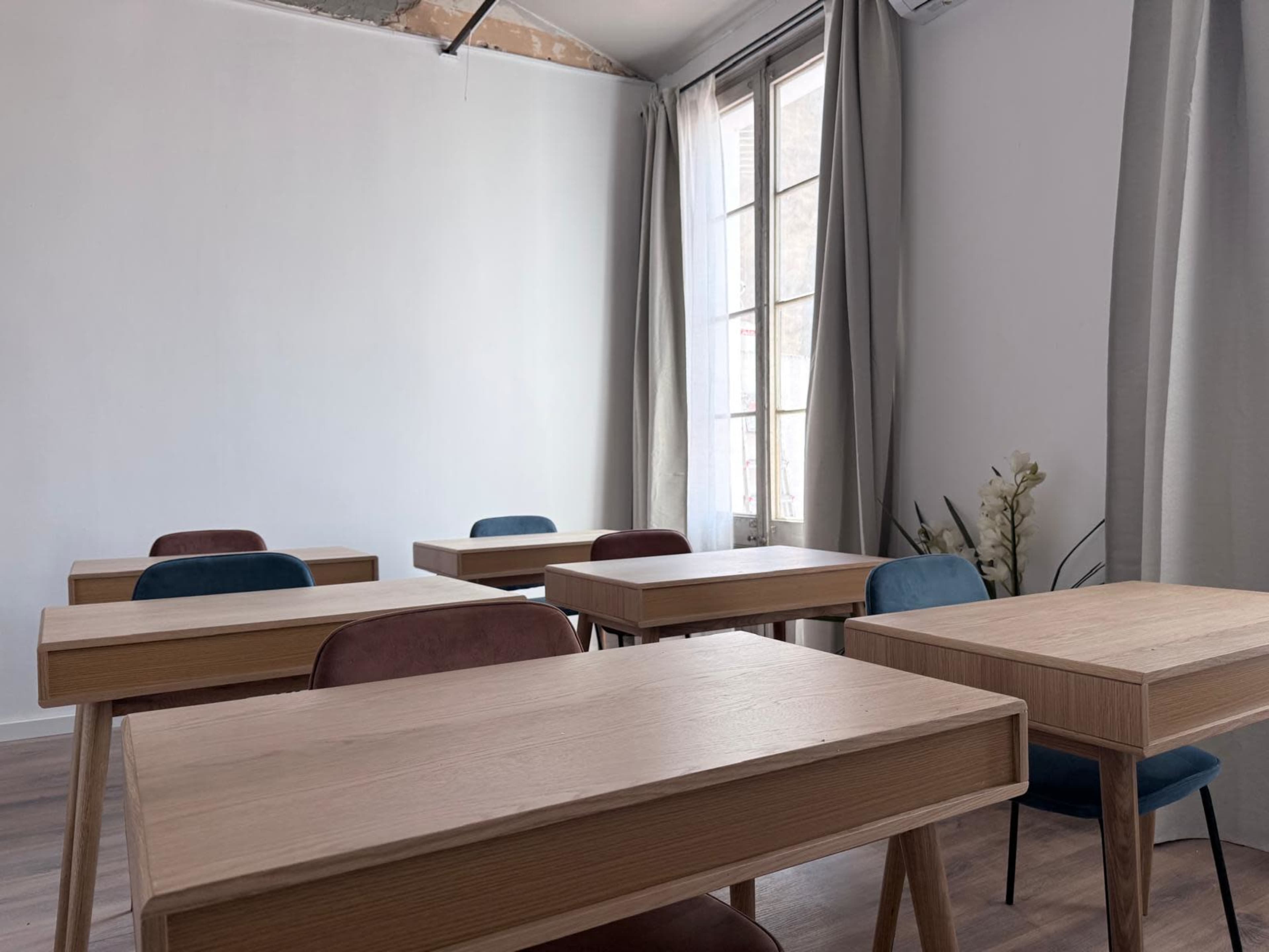 The image shows a minimalist classroom with wooden desks arranged in rows and large windows covered by light curtains.