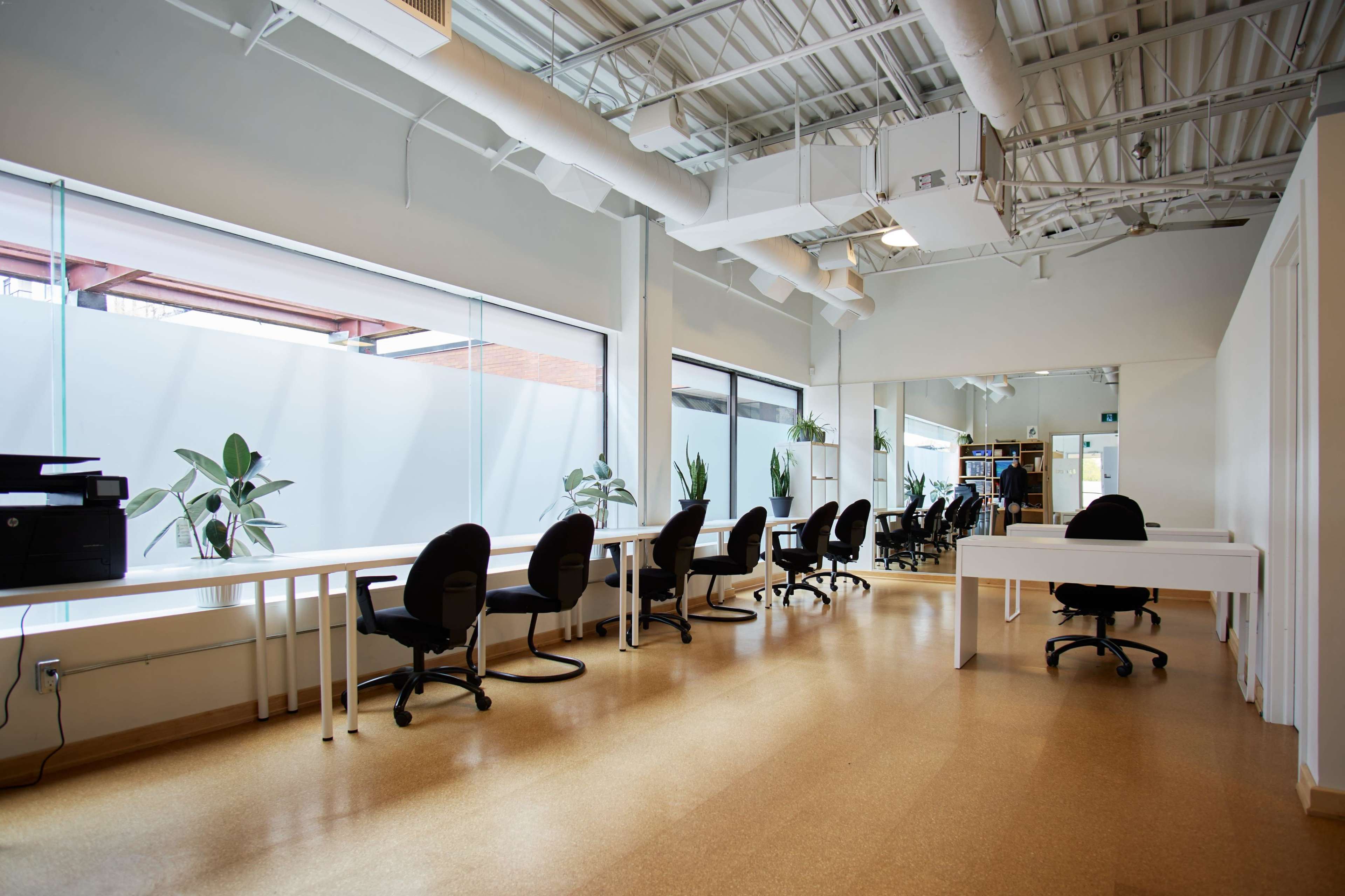 A modern office space with large windows, several desks and chairs, and potted plants along the walls.
