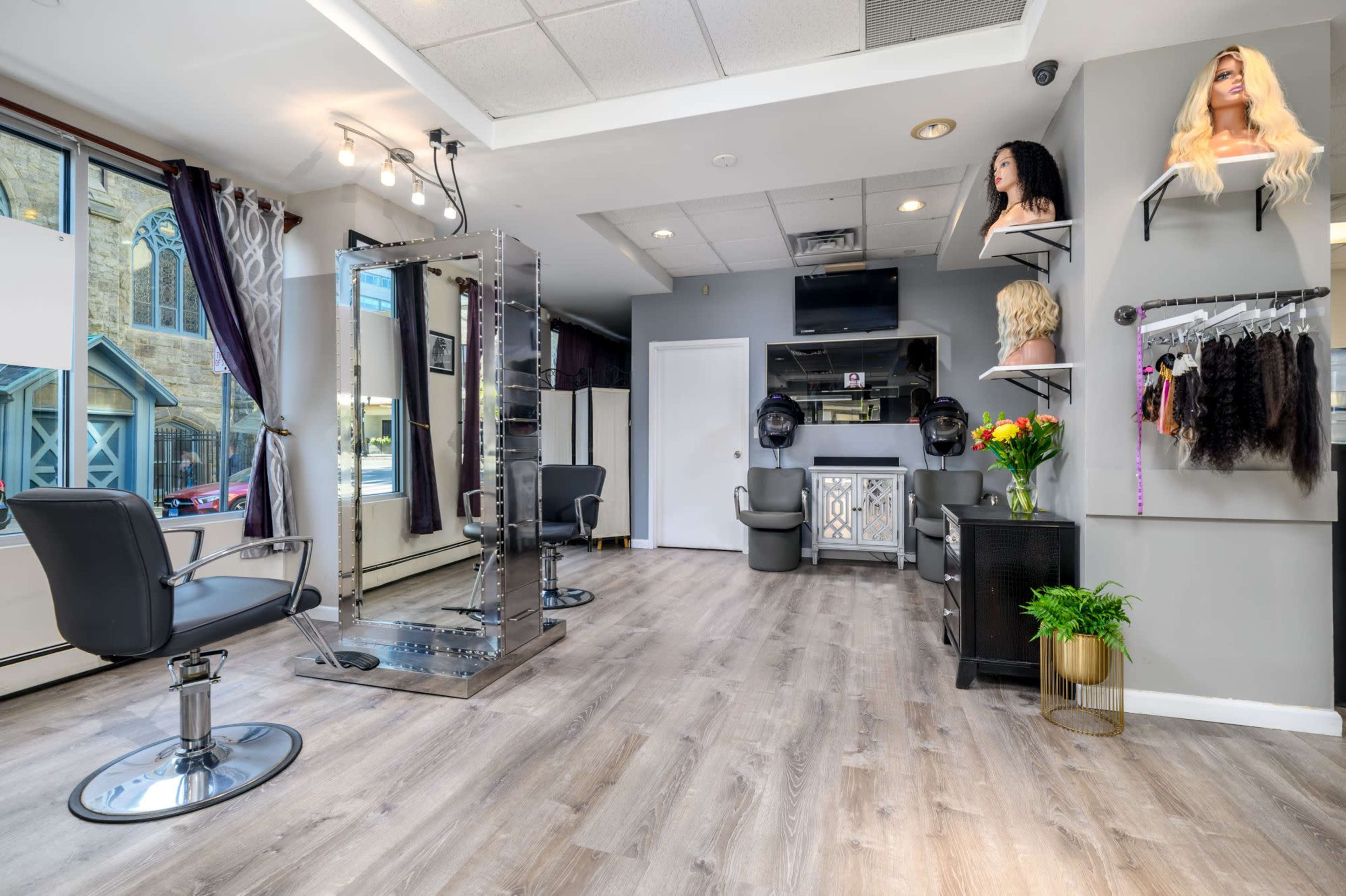 A modern hair salon featuring two styling chairs, a display of wigs on mannequins, and a wall-mounted rack of hair extensions.