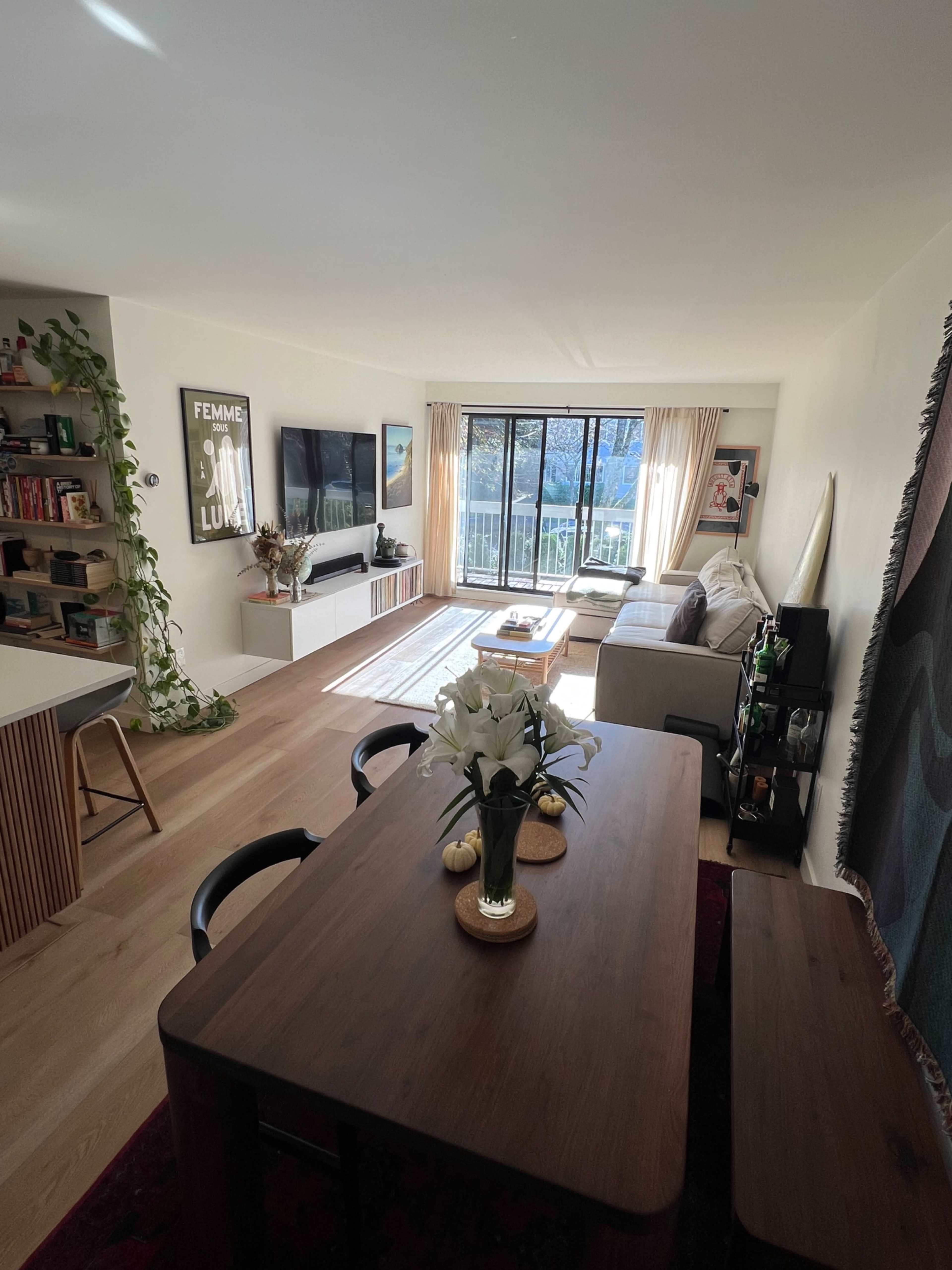 A modern living room and dining area with a large window, a dining table with a vase of flowers, a couch, and a television mounted on the wall.