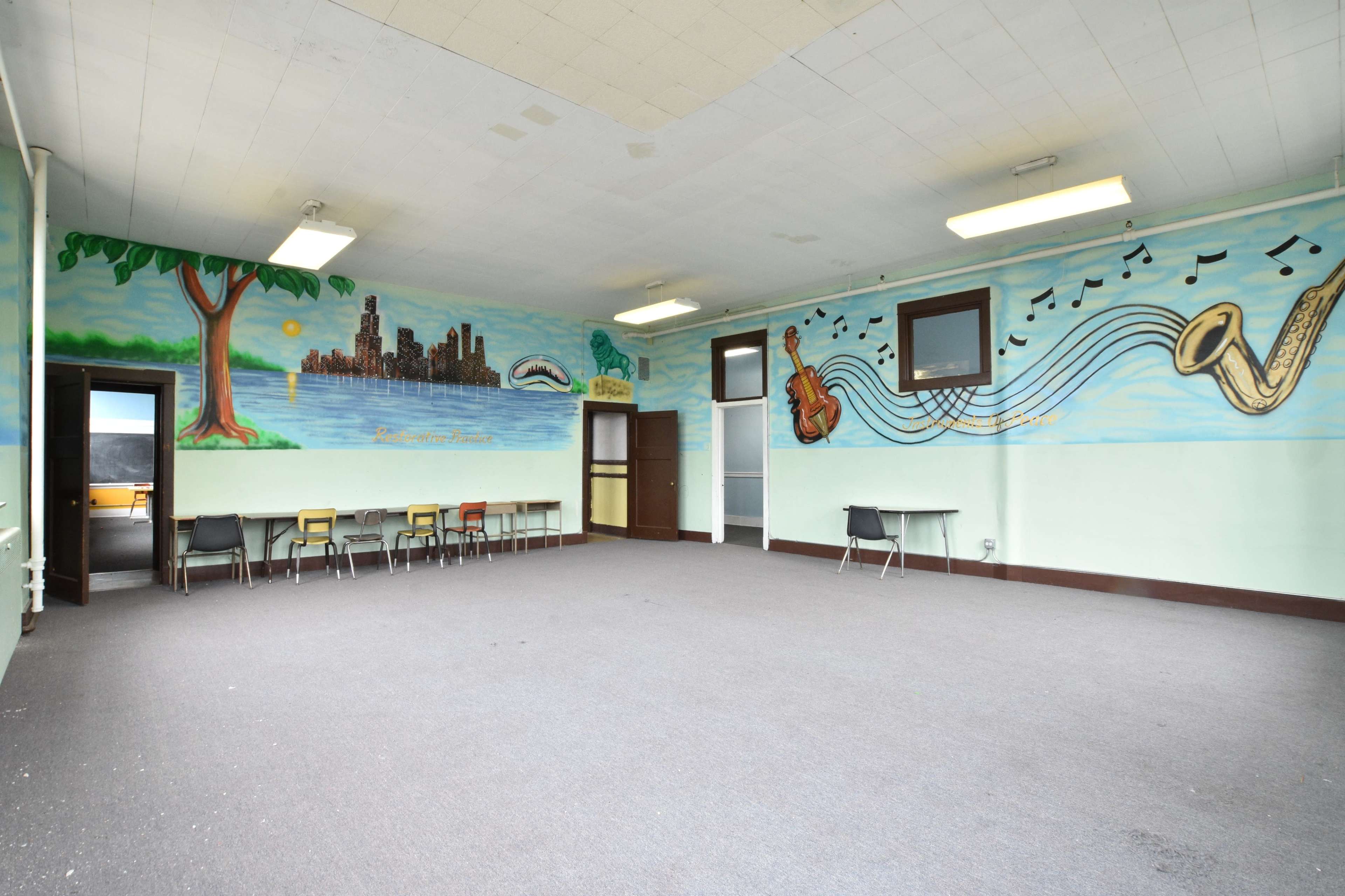 An empty room features large murals on the walls depicting a city skyline, a tree, and musical notes, with several chairs lined up along one side.