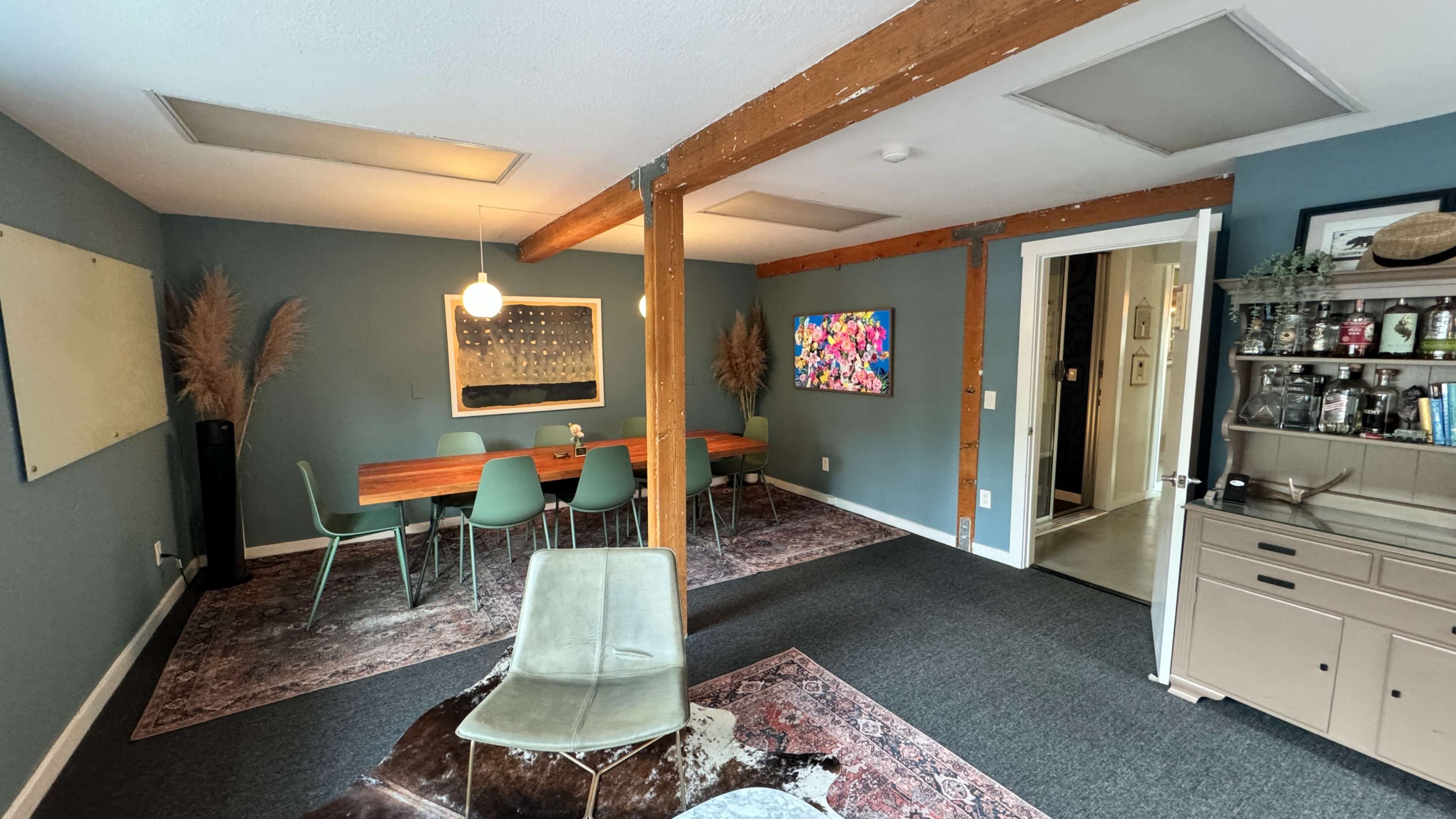 The room features a dining area with a wooden table surrounded by green chairs, a seating area with a chair on a patterned rug, and two doors leading to adjacent spaces.