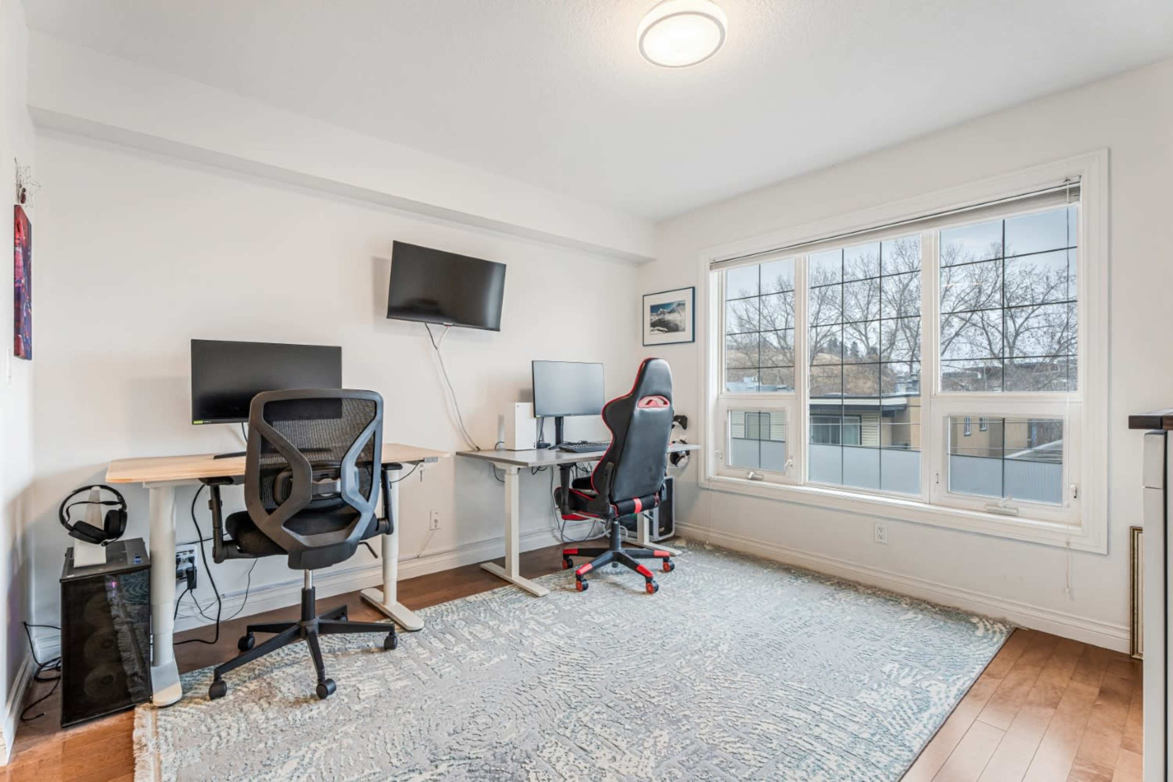 The image shows a modern home office space featuring two desks with computers, two ergonomic chairs, and a window offering natural light.