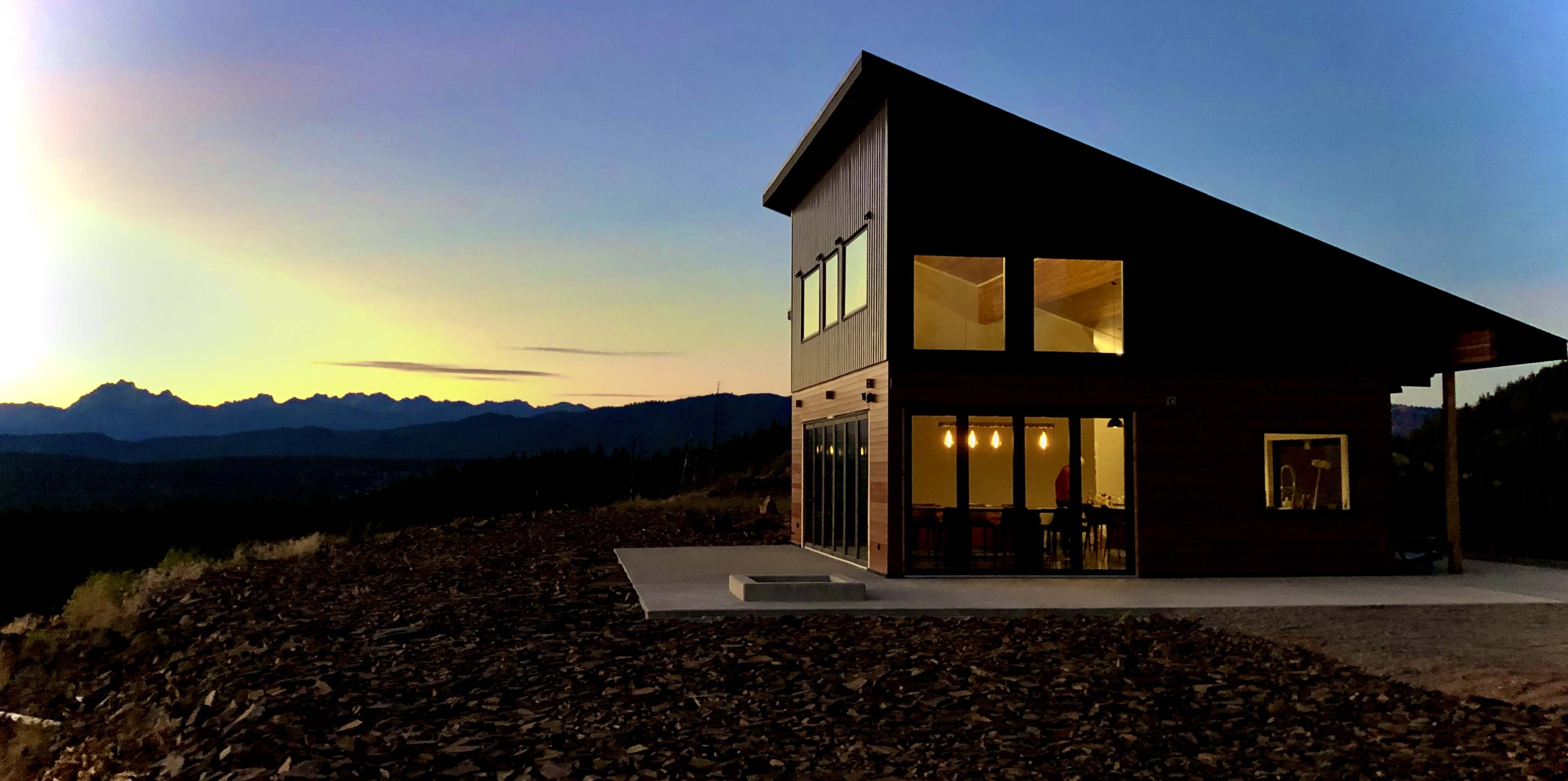 A modern house with large windows sits on a rocky terrain, silhouetted against a colorful sunset over distant mountains.