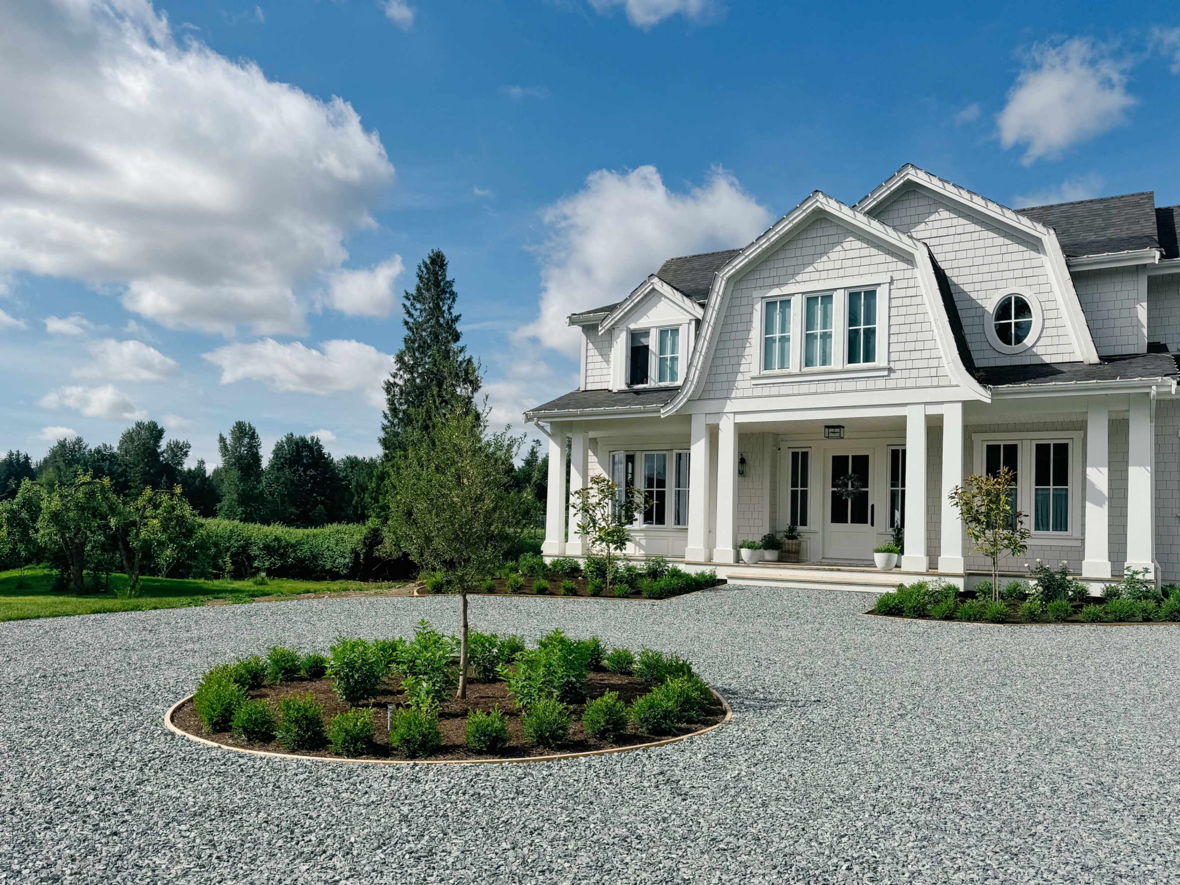 A large white house with a landscaped front yard and gravel driveway is shown under a partly cloudy sky.