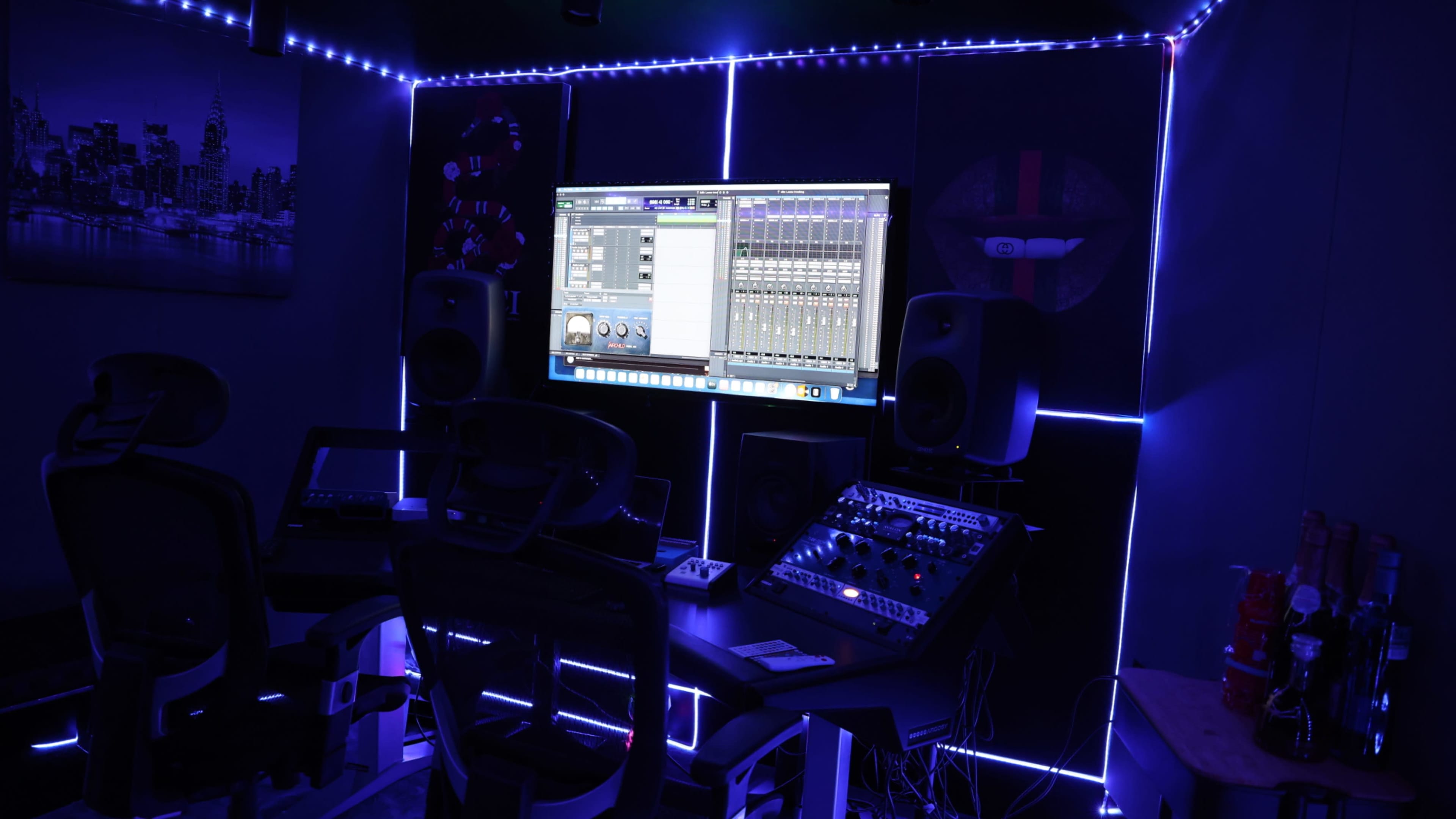 A music production studio featuring a large monitor displaying audio editing software, surrounded by sound equipment and illuminated with blue LED lights.