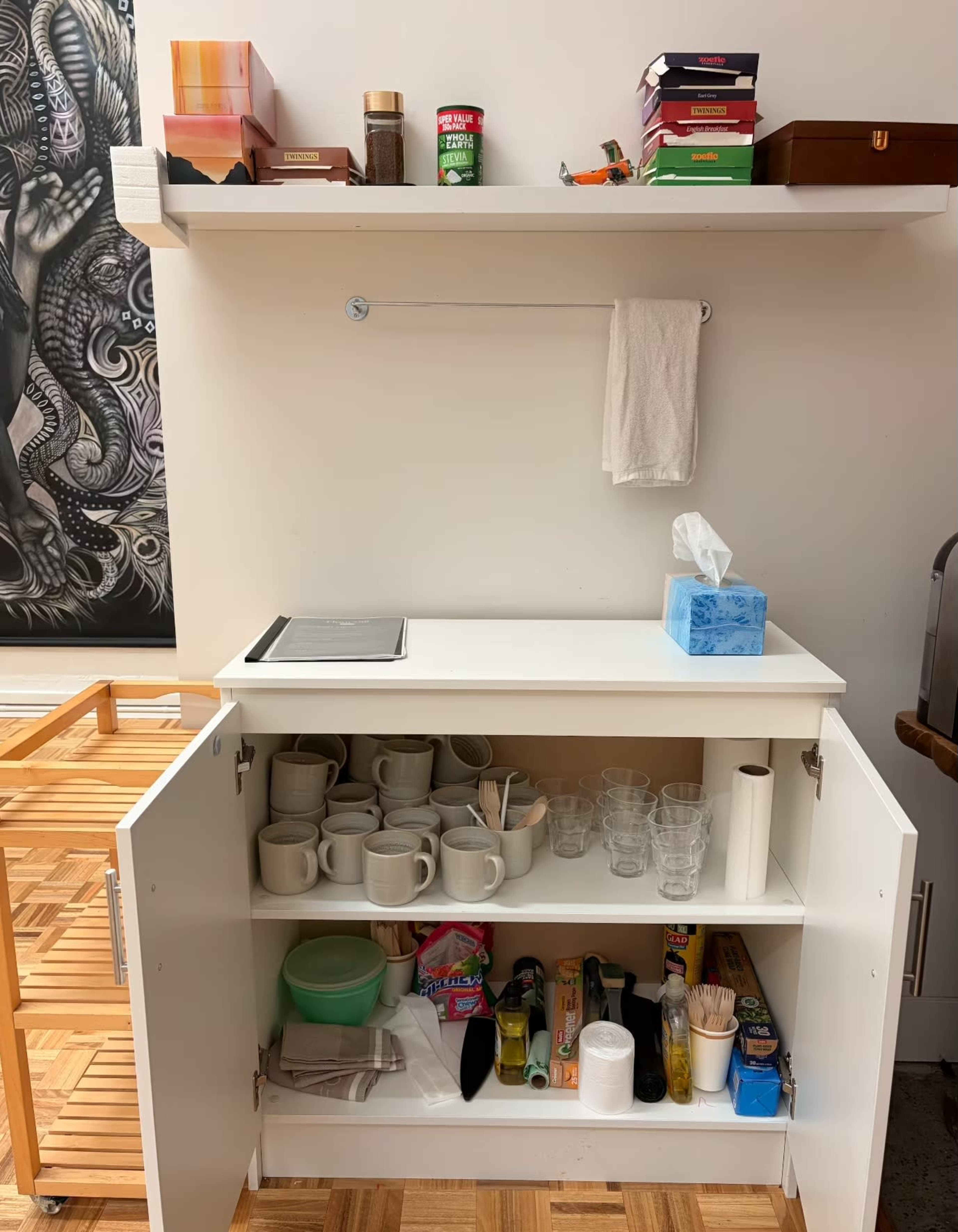 The image shows a white cupboard with a variety of mugs, glasses, and utensils stored on shelves, along with a small countertop and items on the wall above.