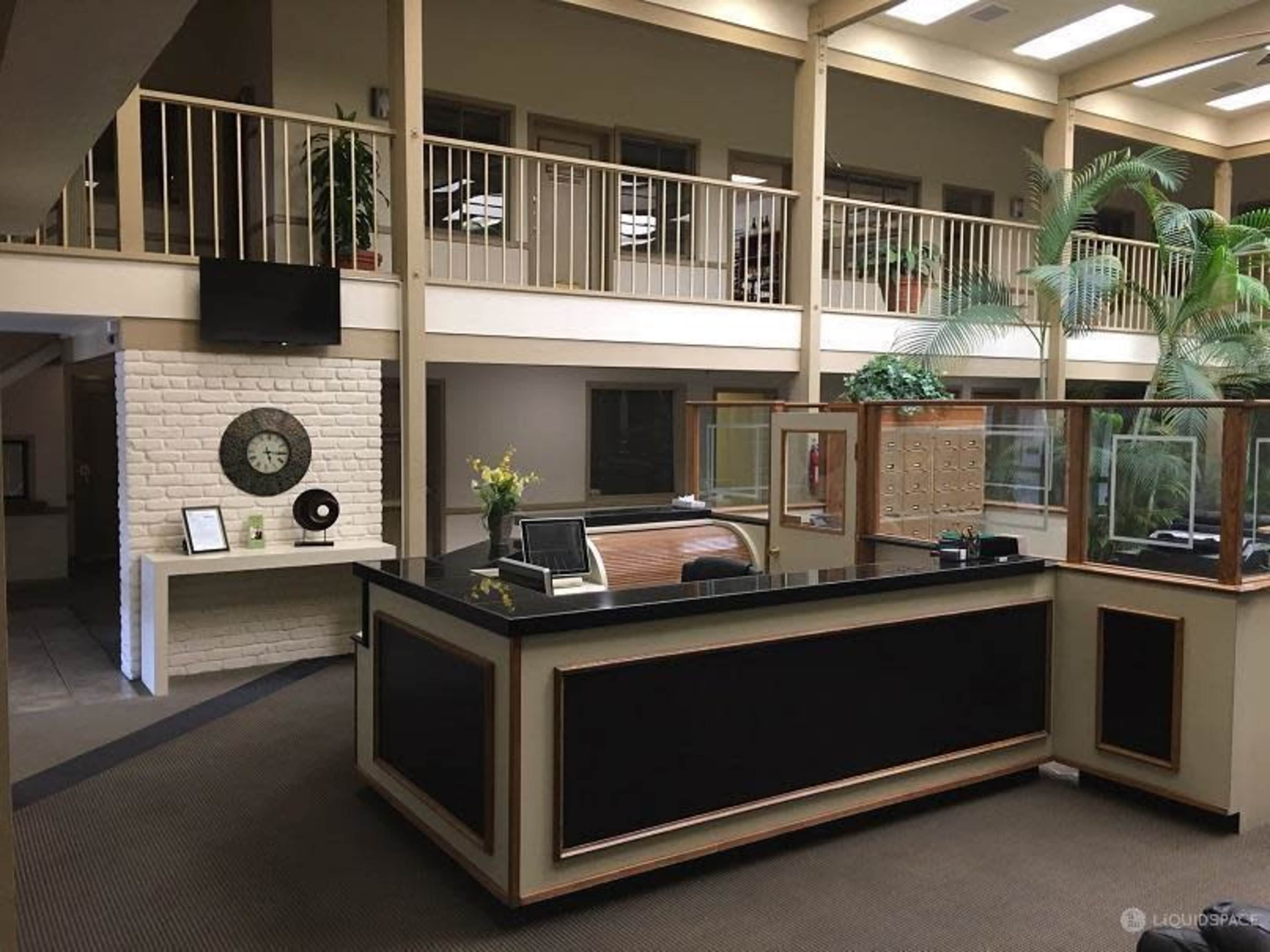 The image shows a reception area with a black front desk, a decorative plant, and a two-story layout featuring a balcony and a light-colored brick wall.