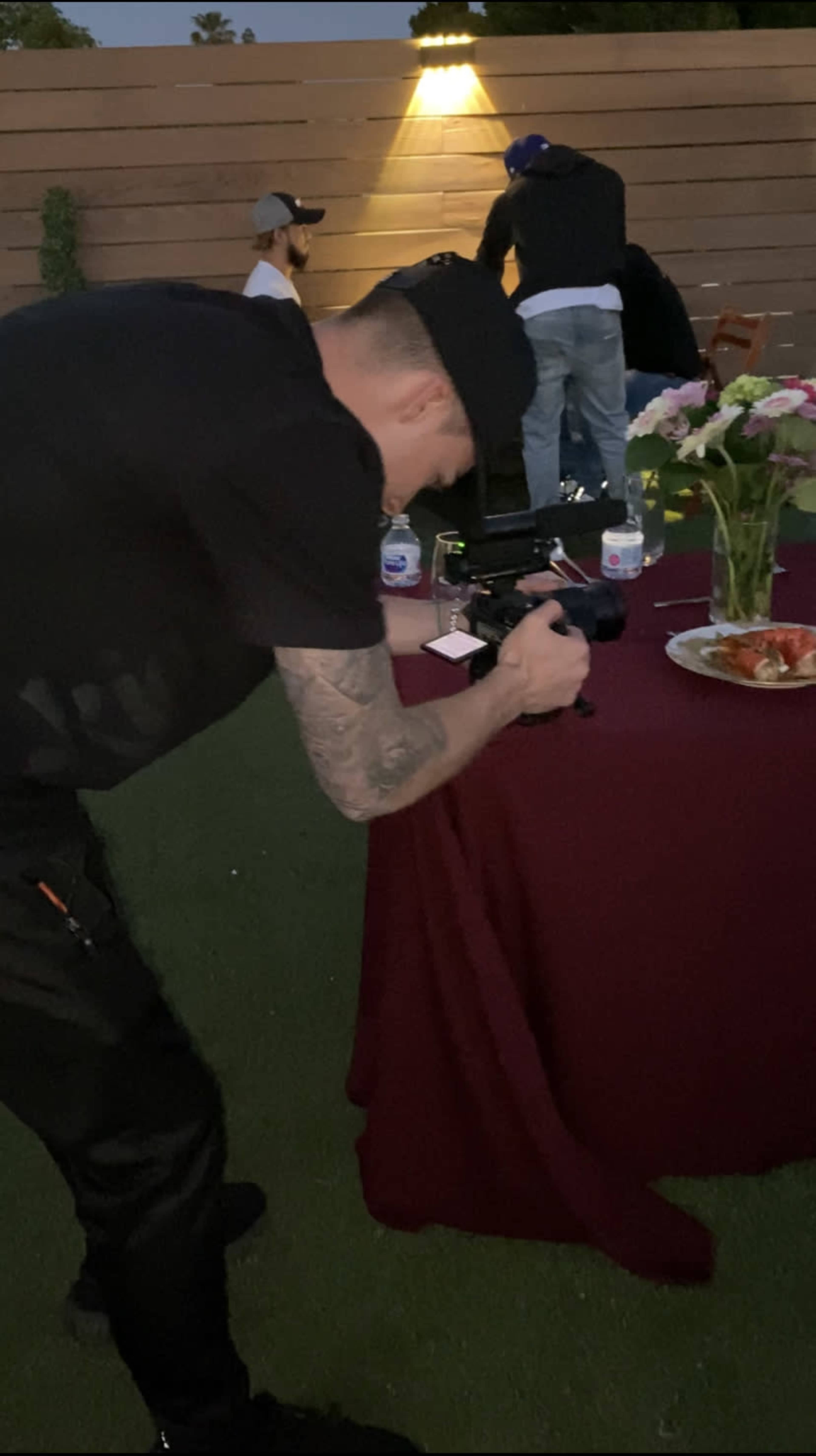 A person with a camera is kneeling in front of a table covered with a red cloth and a plate of food, while others are gathered in the background.