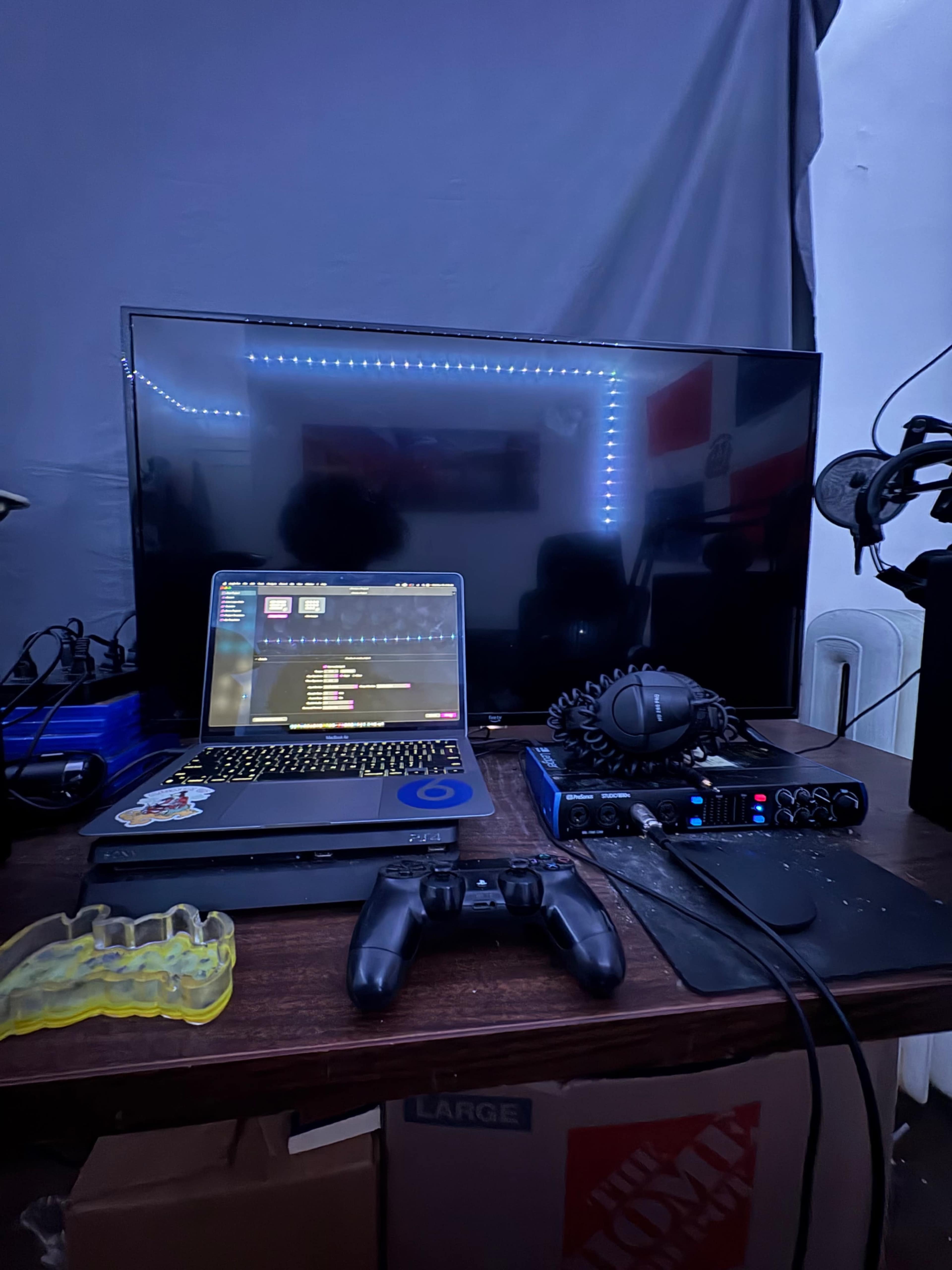 A laptop displaying code sits on a desk beside a large television, a gaming controller, a set of headphones, and an audio interface, all in a dimly lit room.