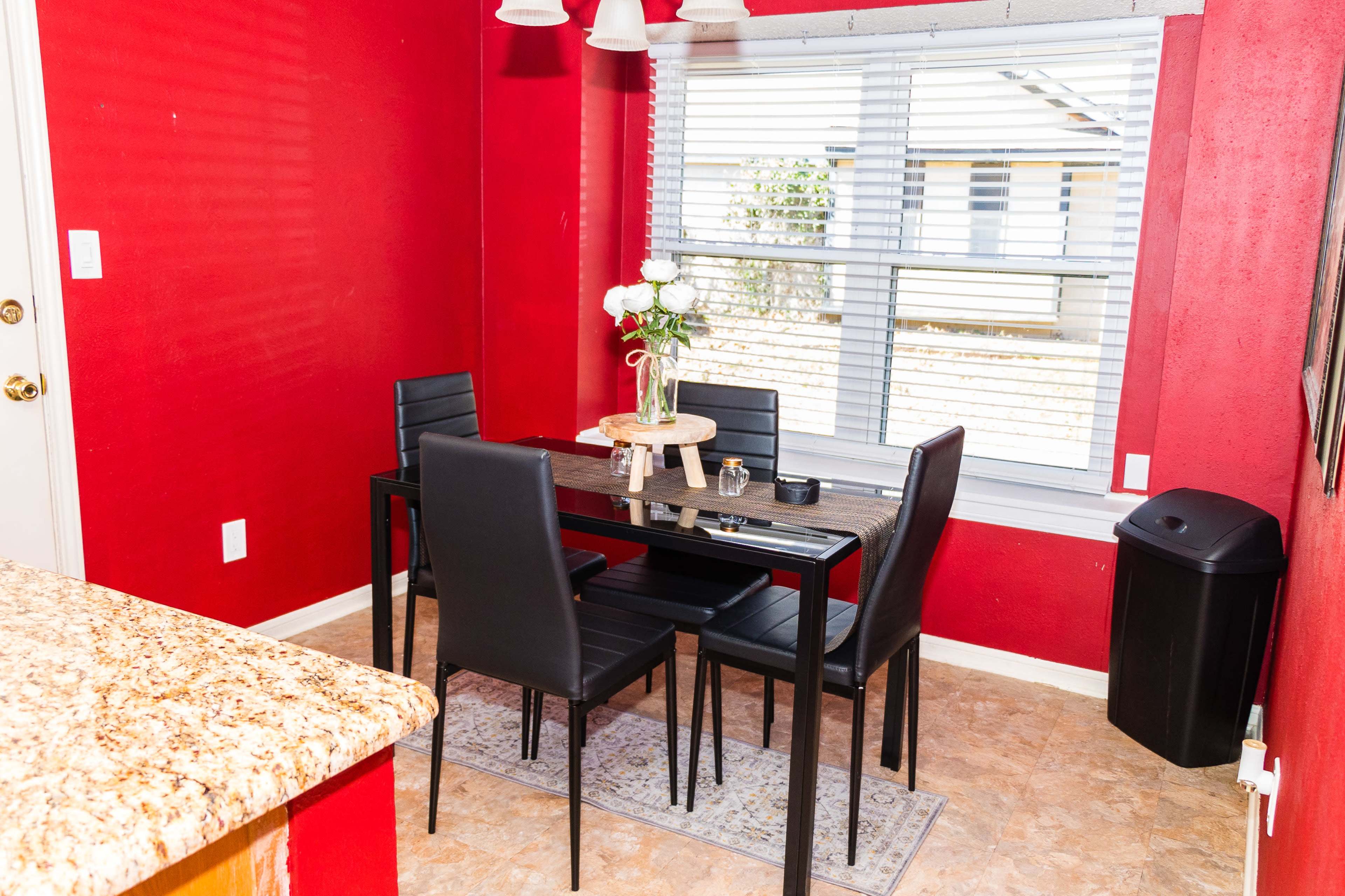 A dining area features a black table with four chairs, a vase of flowers, and a window allowing natural light, all set against a red wall.
