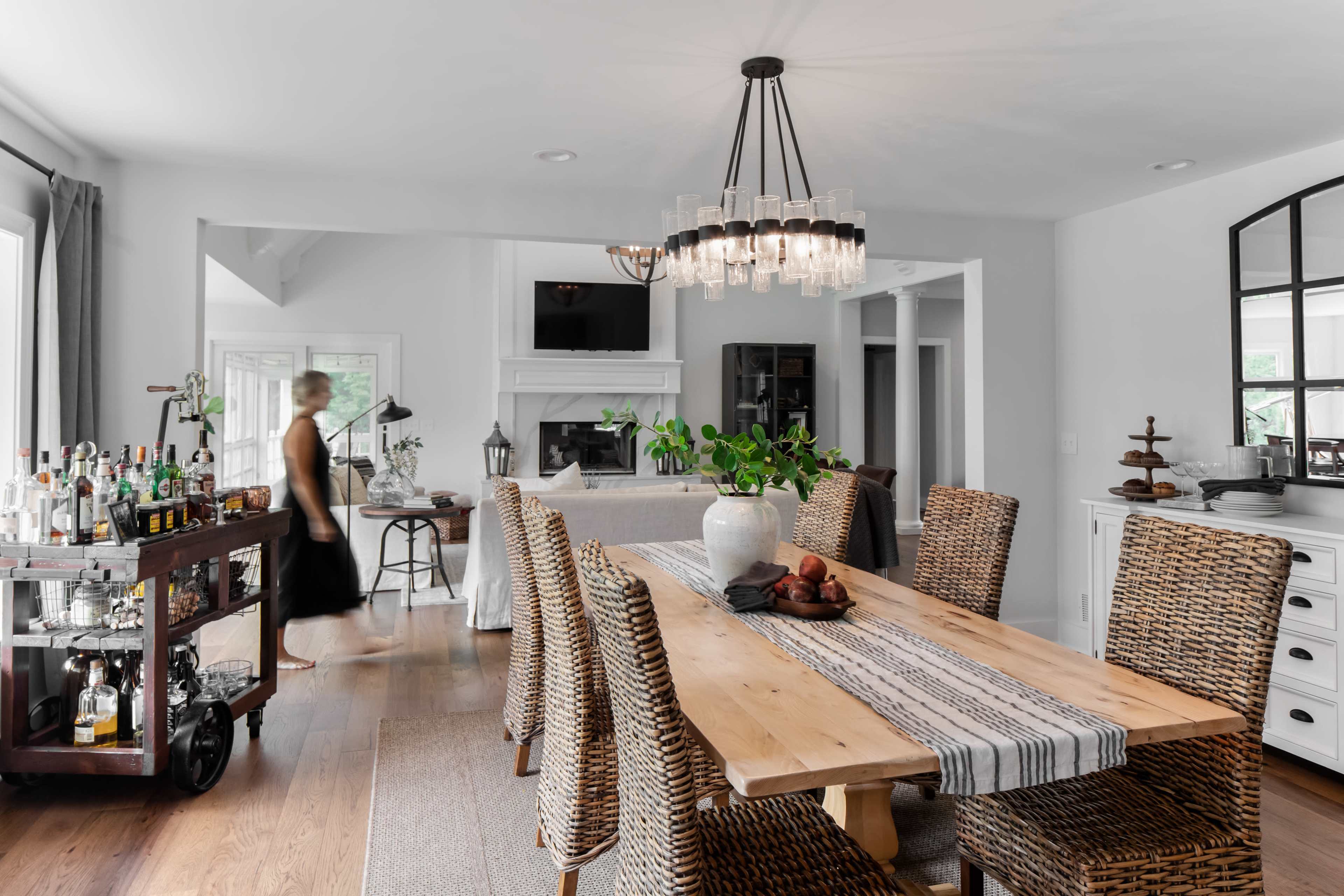 A dining area features a large wooden table surrounded by woven chairs, with a bar cart and decorative elements in the background.