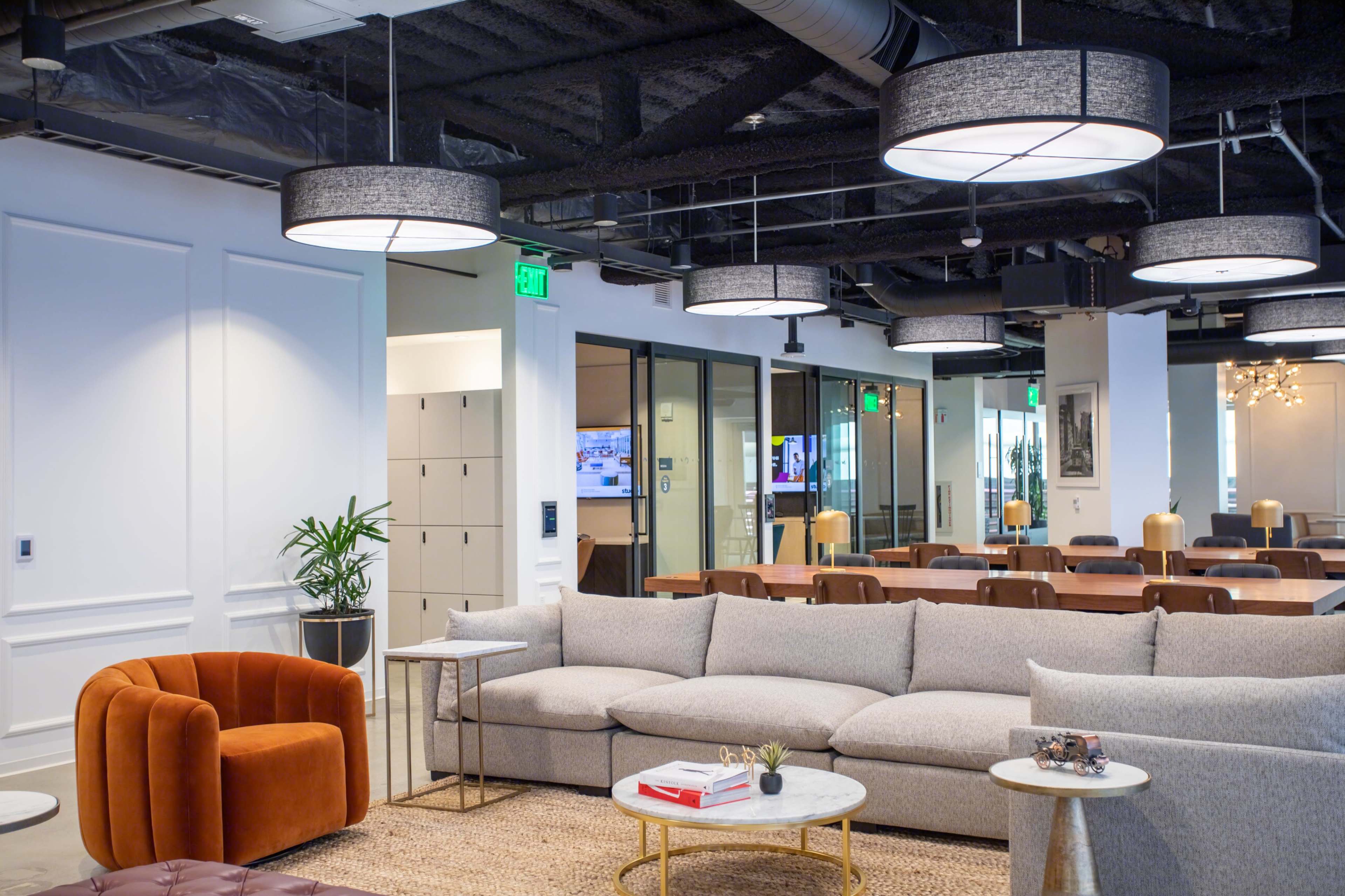 A modern office lounge area featuring a large gray sectional sofa, an orange accent chair, and contemporary pendant lights, with a dining area visible in the background.