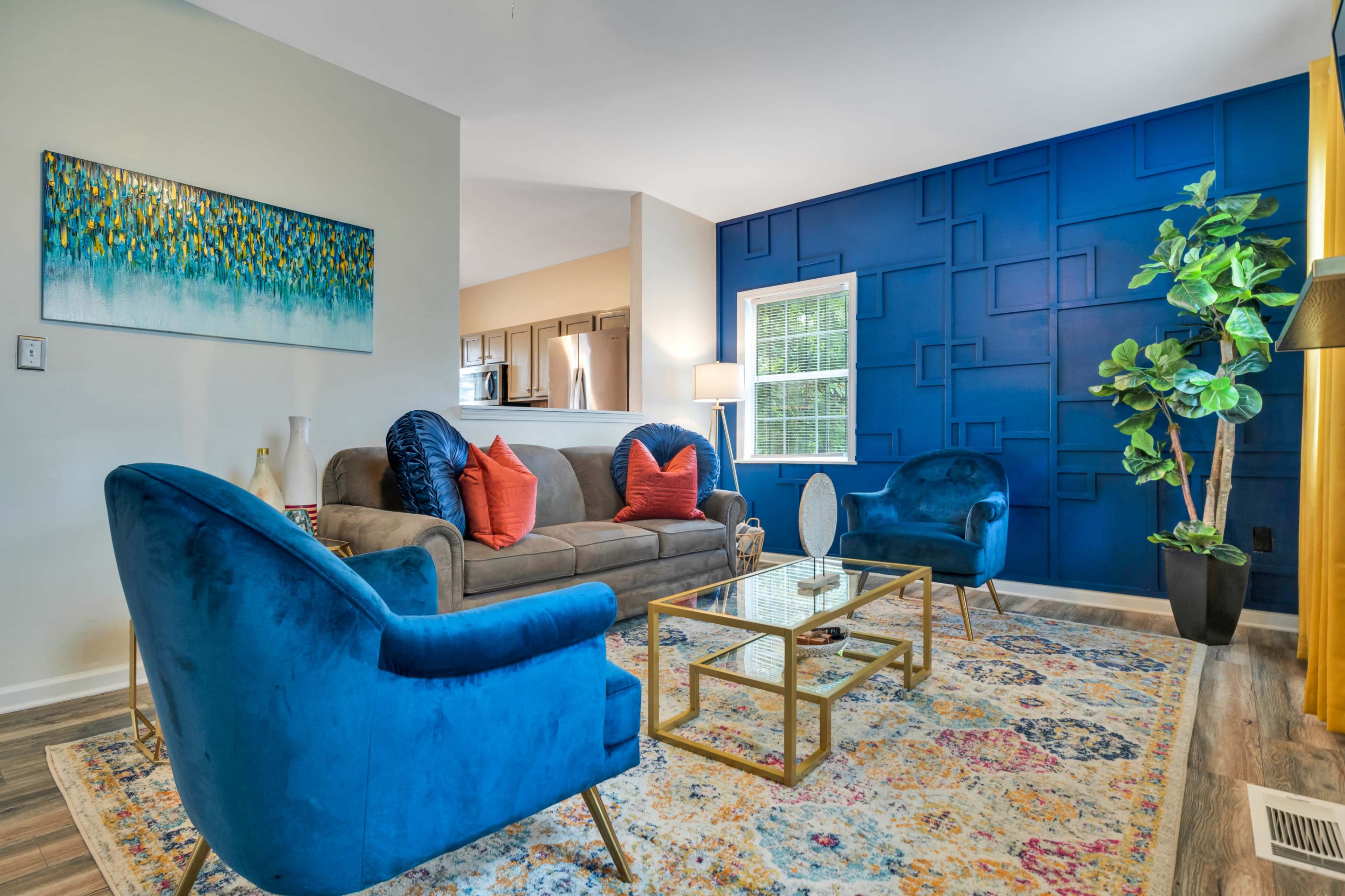 A living room featuring a blue accent wall, a gray sofa with orange pillows, two blue armchairs, and a glass coffee table, along with a plant and decorative artwork.