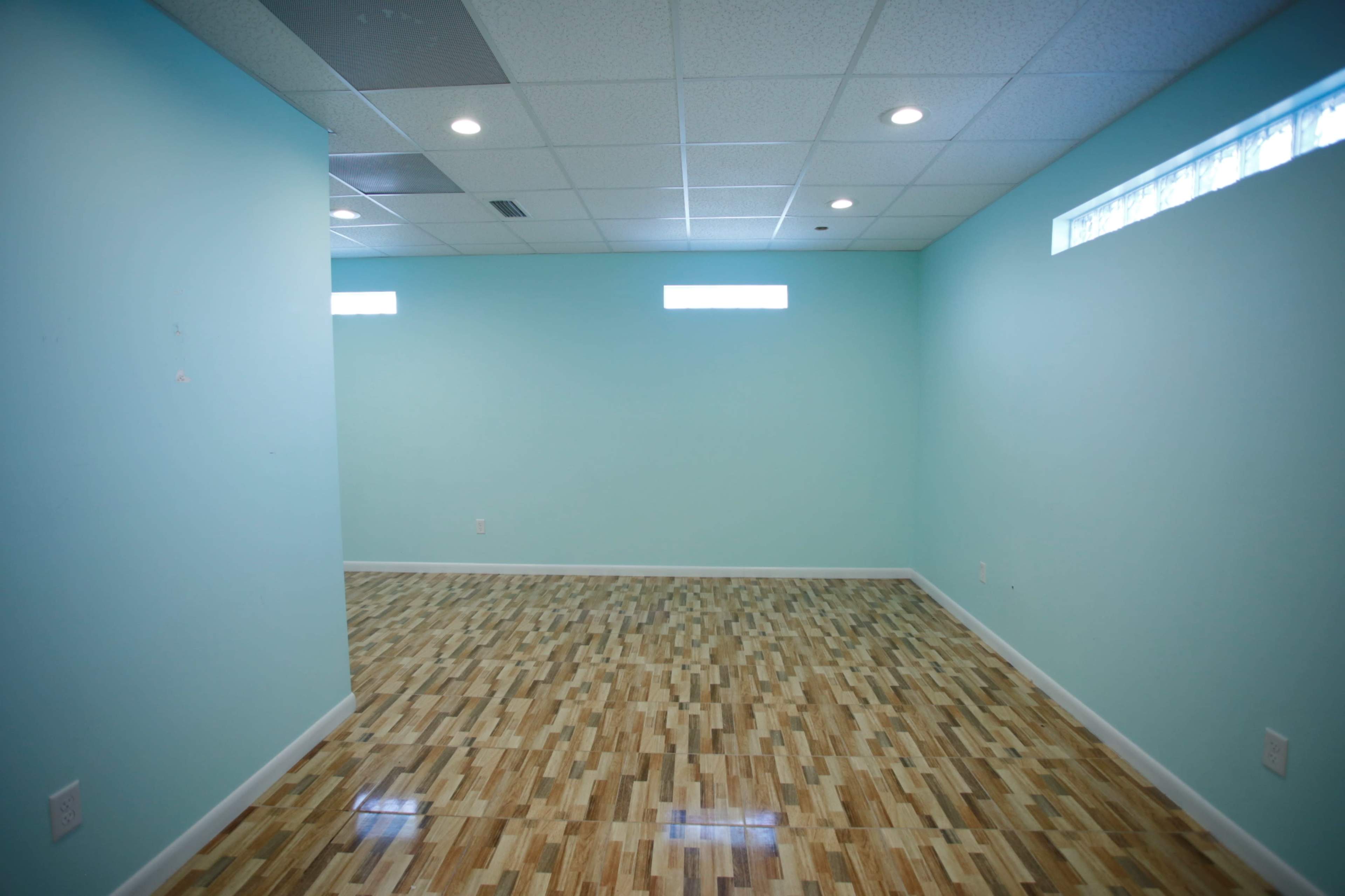 The image shows an empty room with light blue walls, wooden flooring, and small windows on one side.