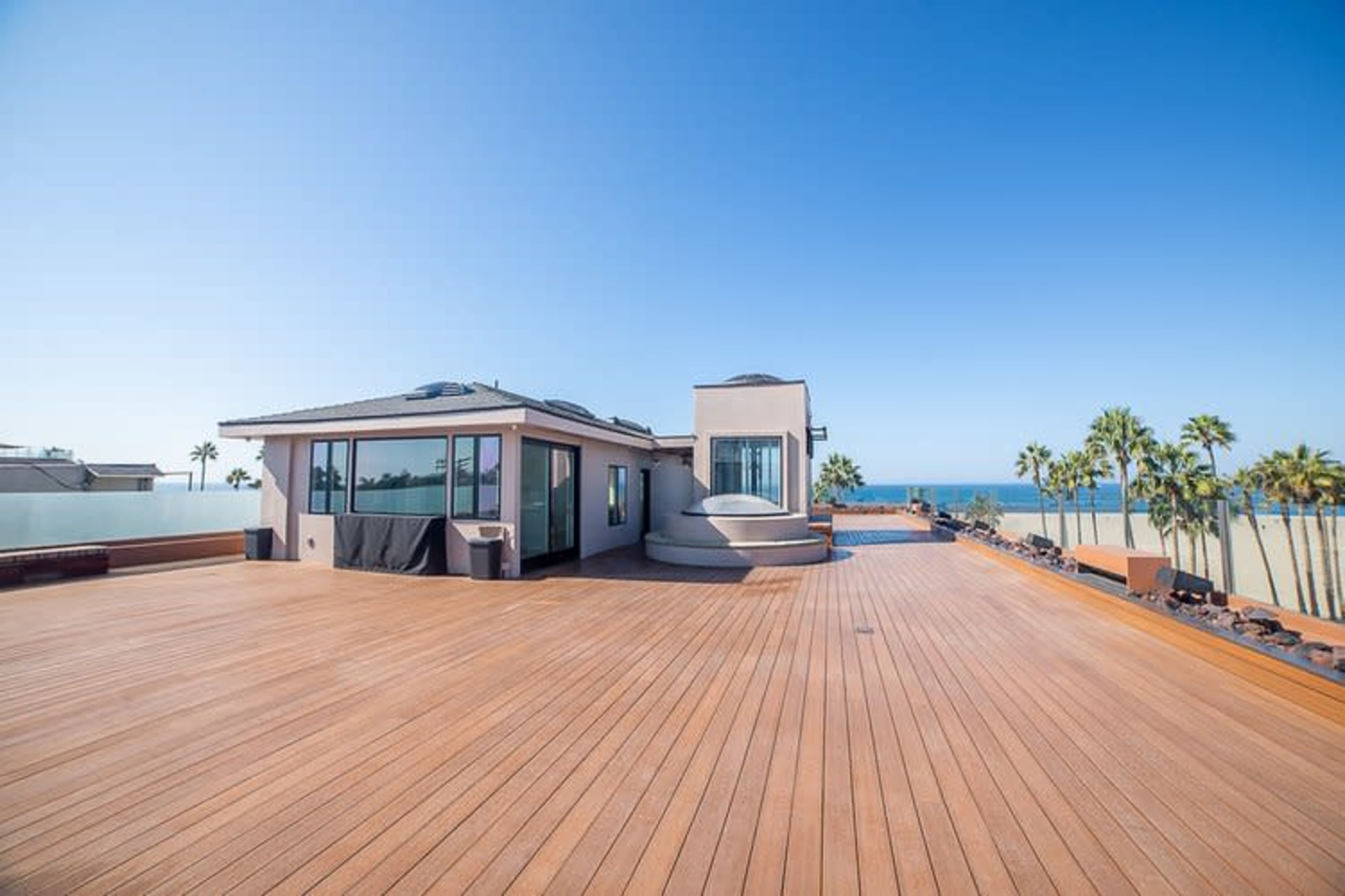 A rooftop deck features a spacious wooden surface with a view of the ocean and palm trees, accompanied by a modern house with large windows.