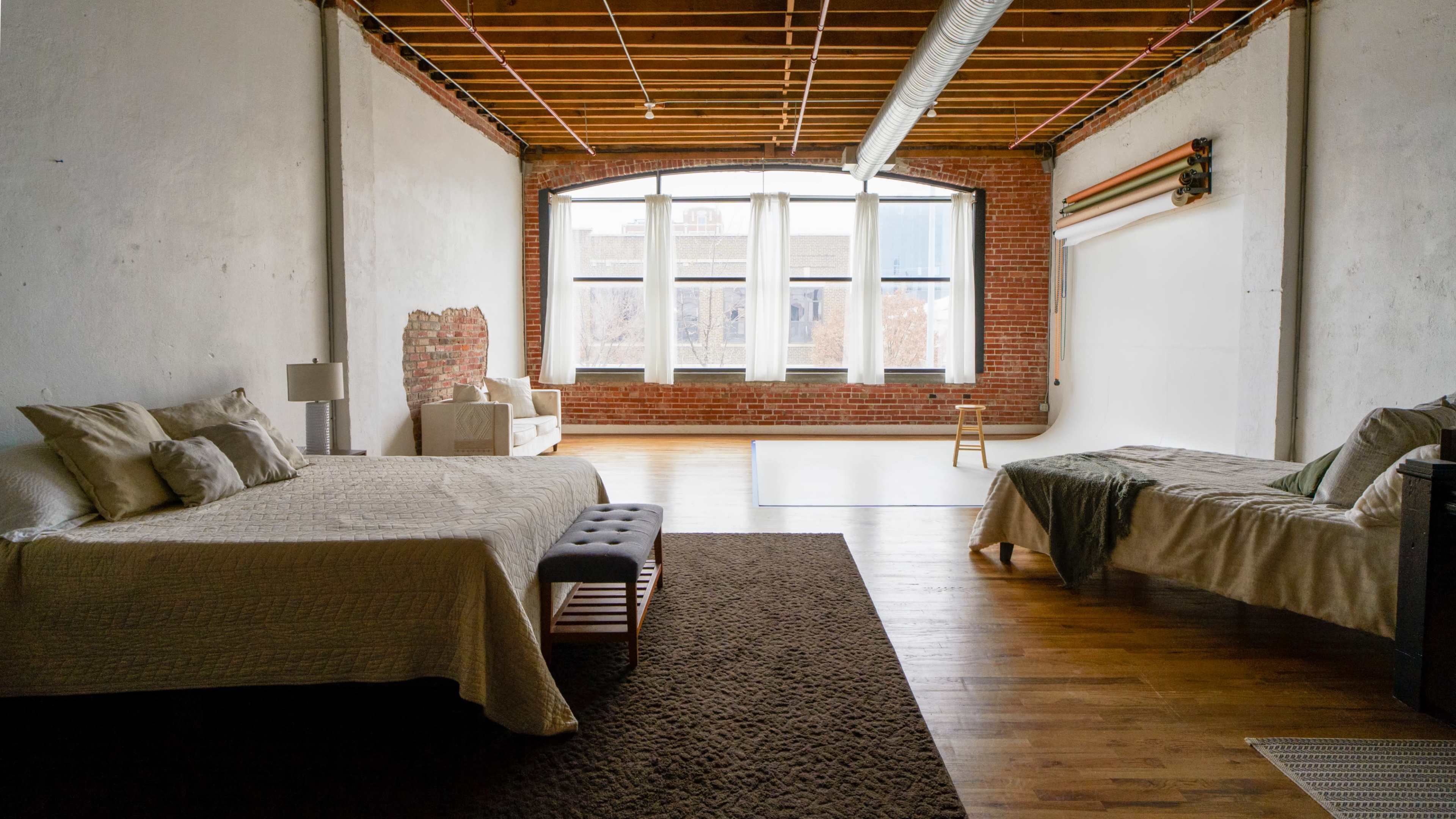A spacious industrial-style room with two beds, large windows, and exposed brick walls.