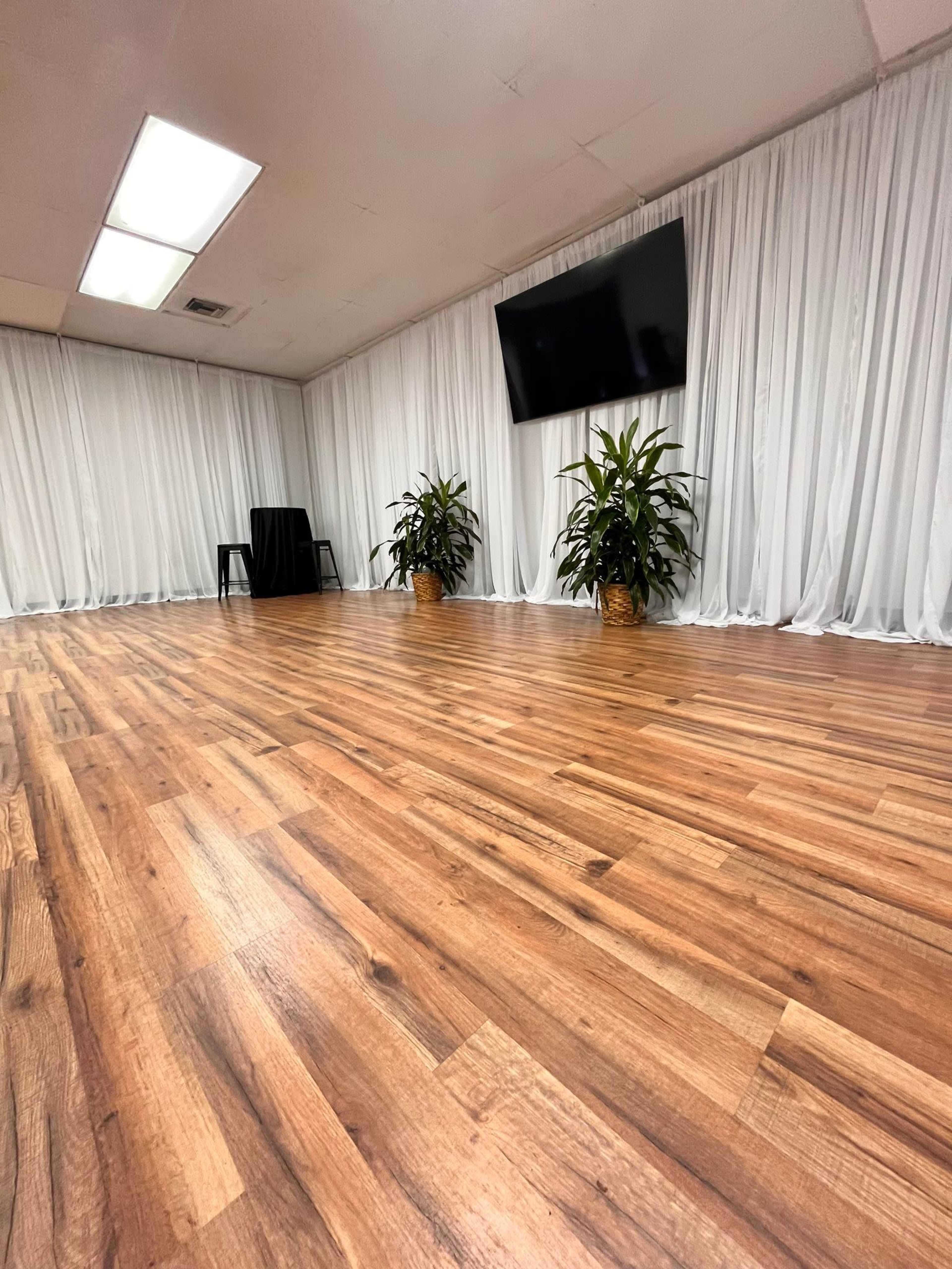 A spacious room features wooden flooring, two potted plants, and a wall-mounted television, with white drapes covering the walls.