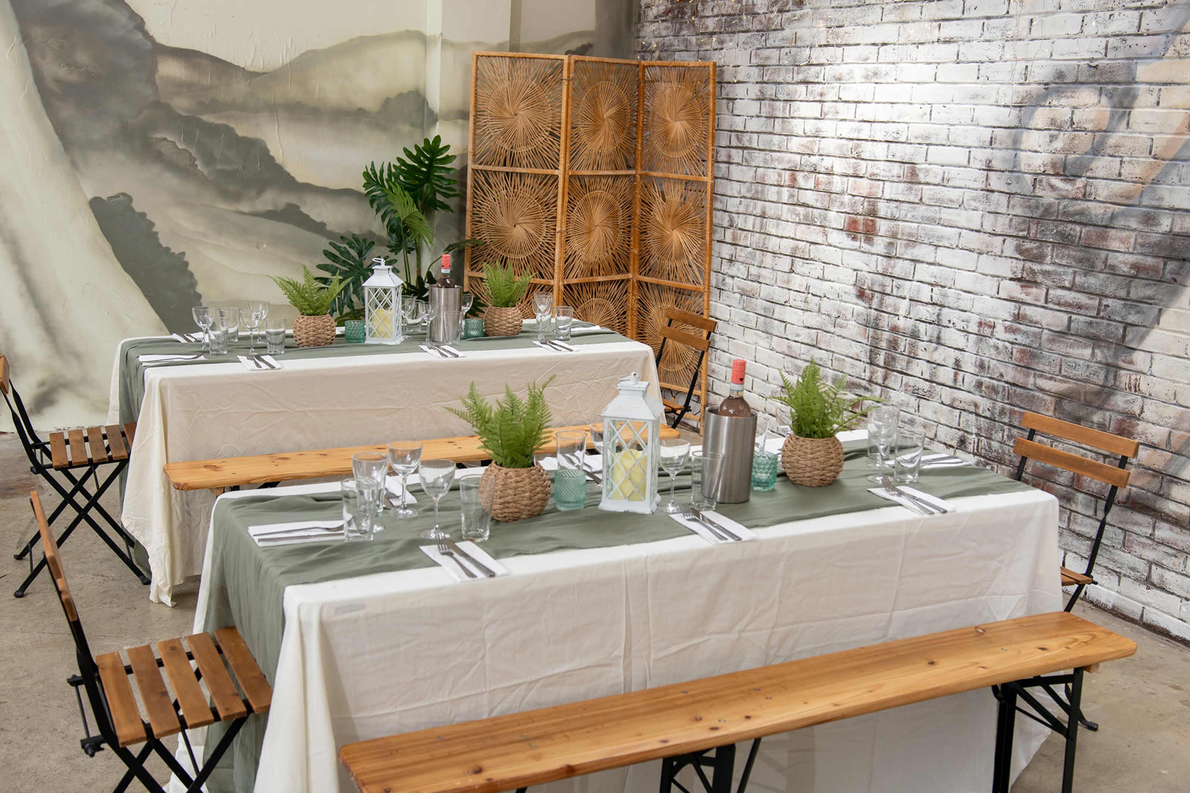 Two tables are set for dining, adorned with tablecloths, glassware, and centerpieces featuring potted plants, against a backdrop of a brick wall and a bamboo screen.