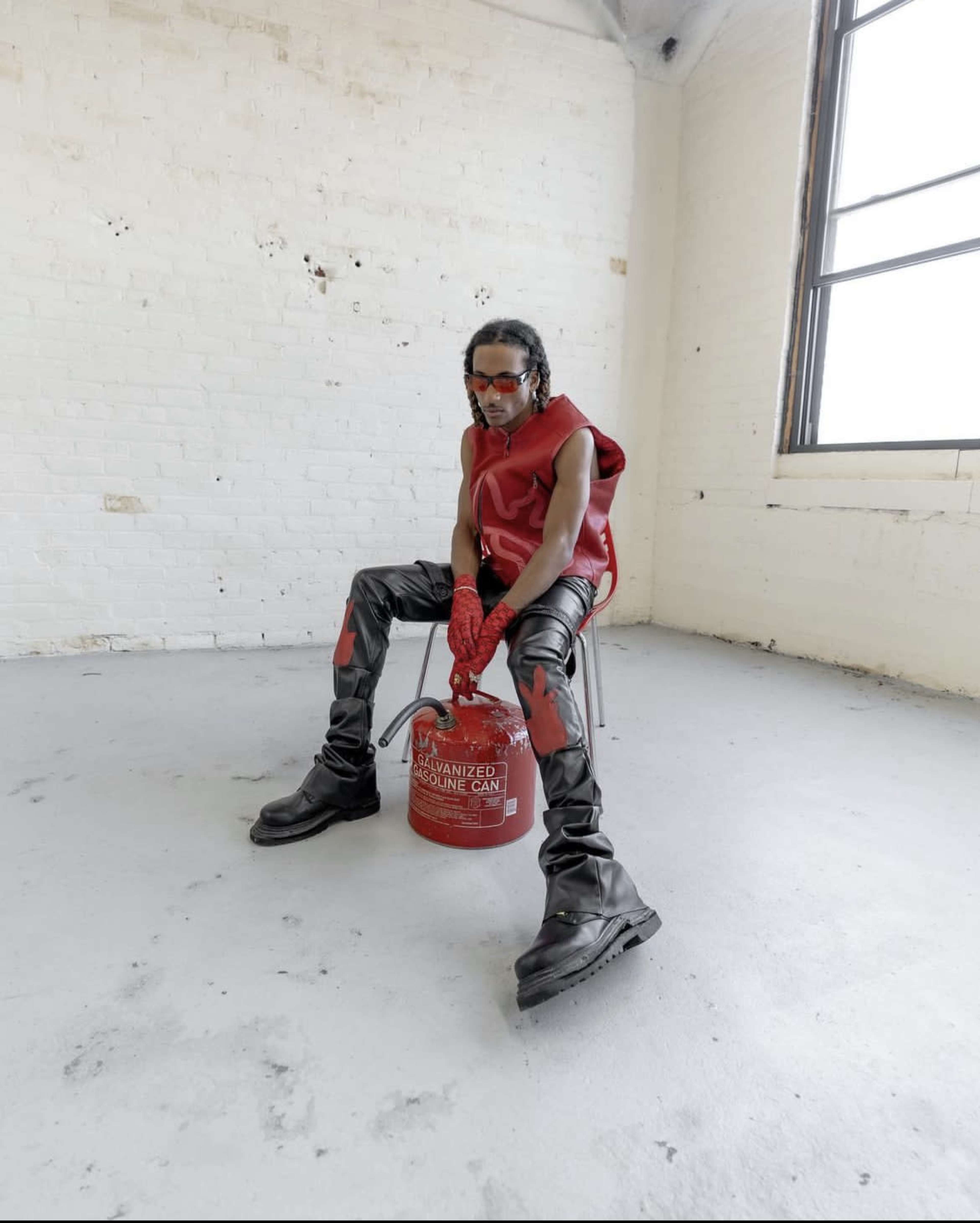A person sits on a chair in an industrial space, wearing red-tinted sunglasses and a red outfit, with black pants and boots, while posing next to a red gasoline can.