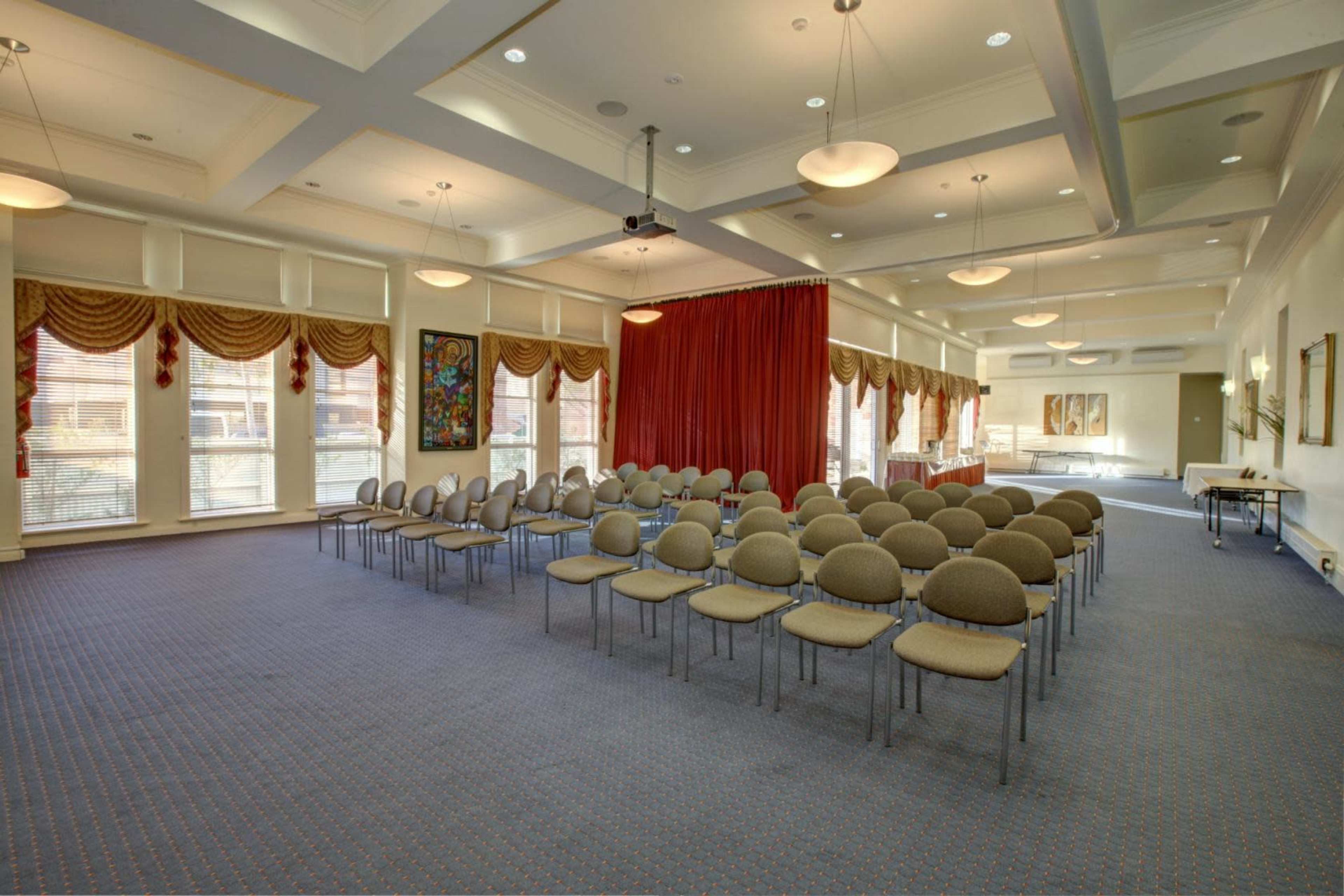 A meeting room set up with rows of circular chairs facing a red curtain, with large windows and decorative drapes along the walls.