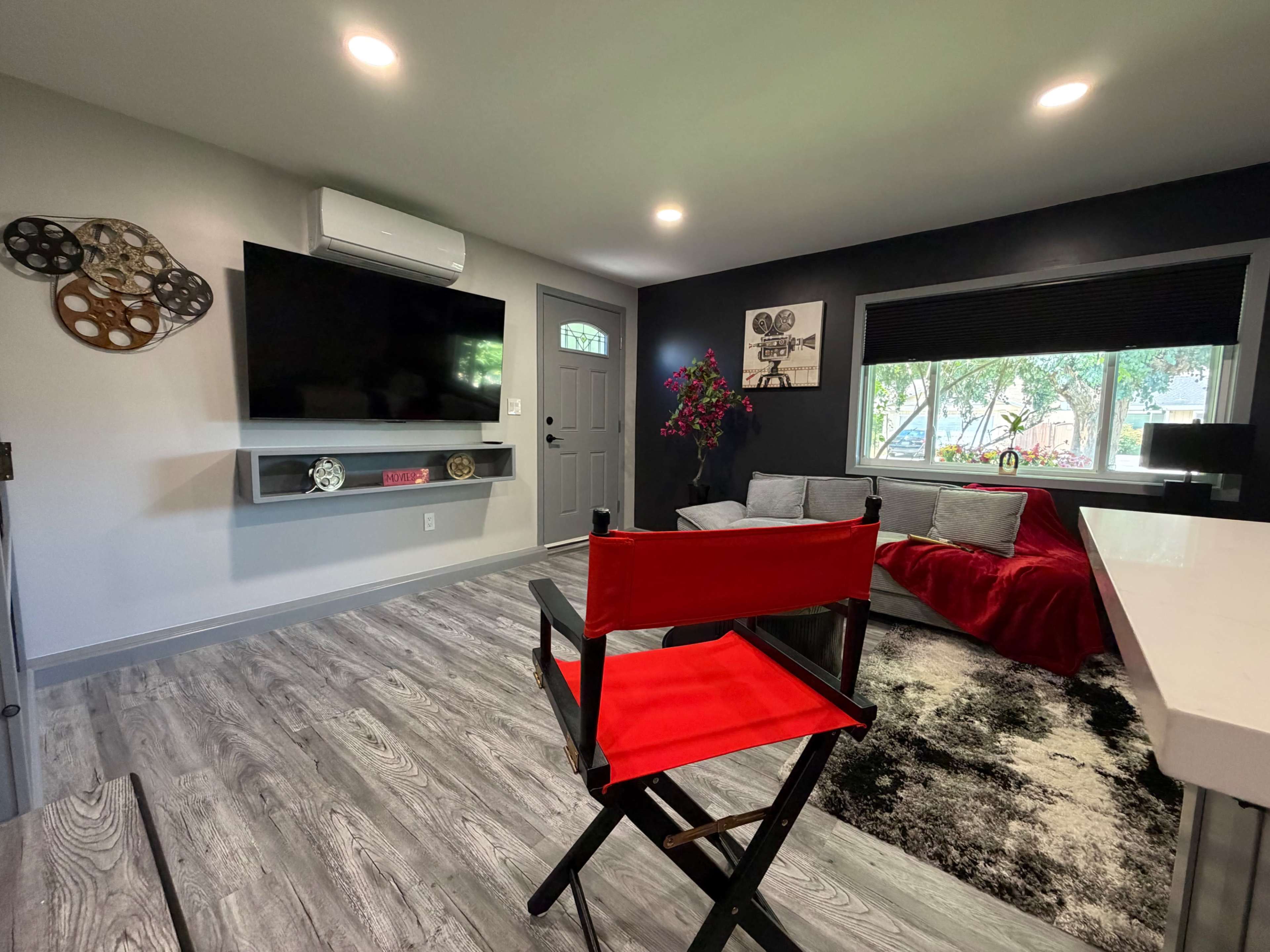 The image shows a modern living room with a black and white color scheme, featuring a large television mounted on the wall, a gray sofa, red throw blanket, and a red director's chair in the foreground.