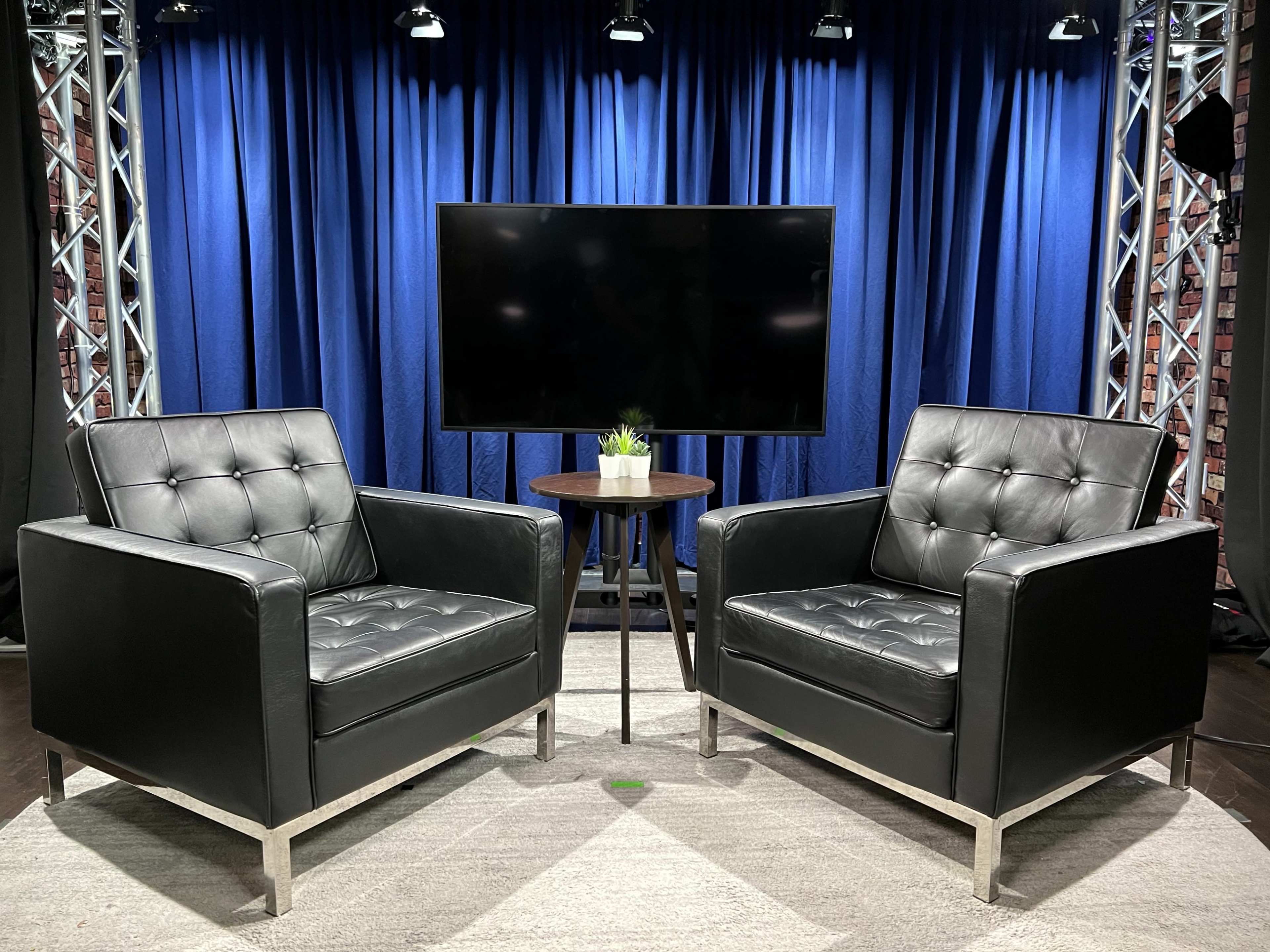 Two black leather armchairs facing each other with a small round table in between, set against a blue curtain backdrop and a large television screen.