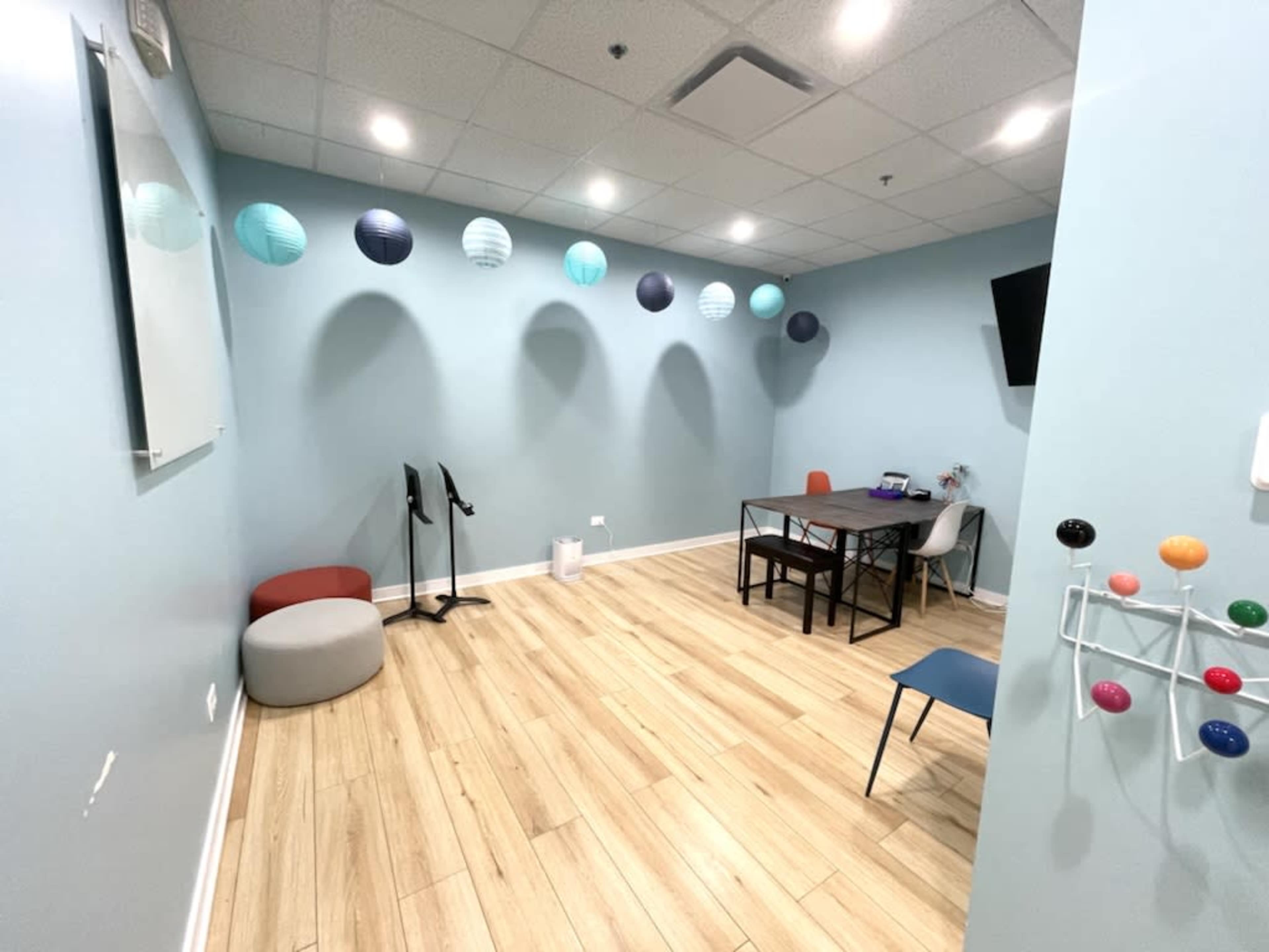 The image shows a light blue room with wooden flooring, featuring a table, chairs, and decorative paper lanterns hanging from the ceiling.