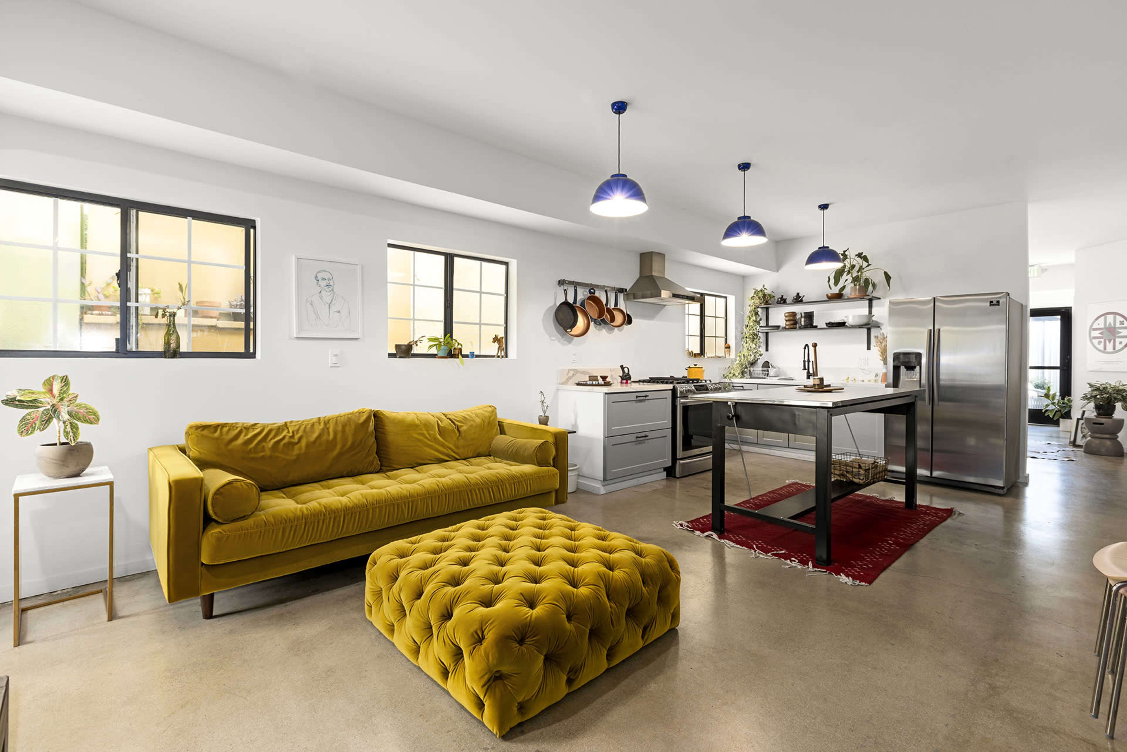 A bright, modern kitchen and living area features a yellow sofa, matching ottoman, and a dining table with industrial-style stools.