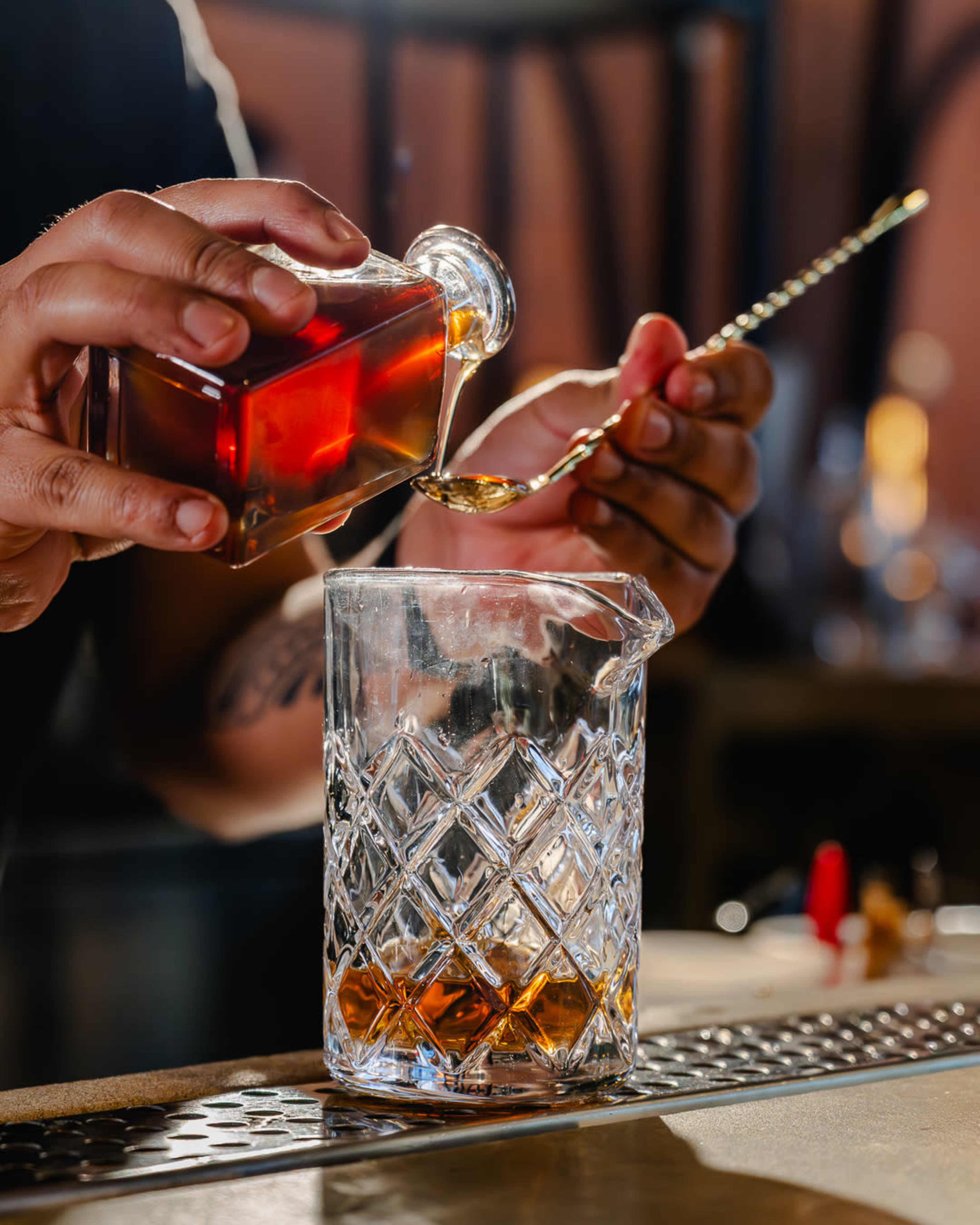 A bartender pours syrup from a glass bottle into a measuring cup on a bar countertop.