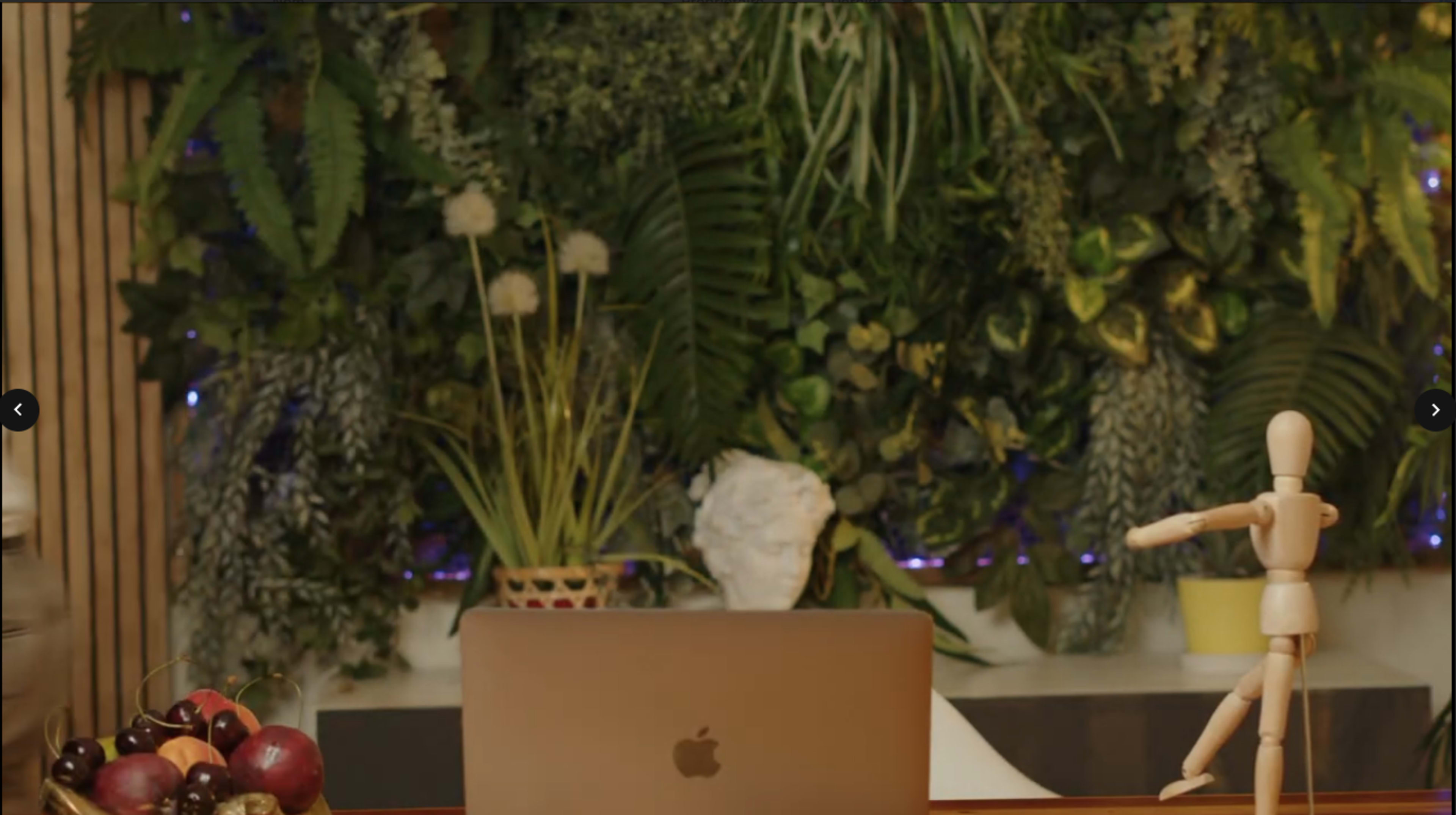 The image features a workspace with a laptop facing a lush vertical garden, complemented by a small potted plant and a wooden mannequin.
